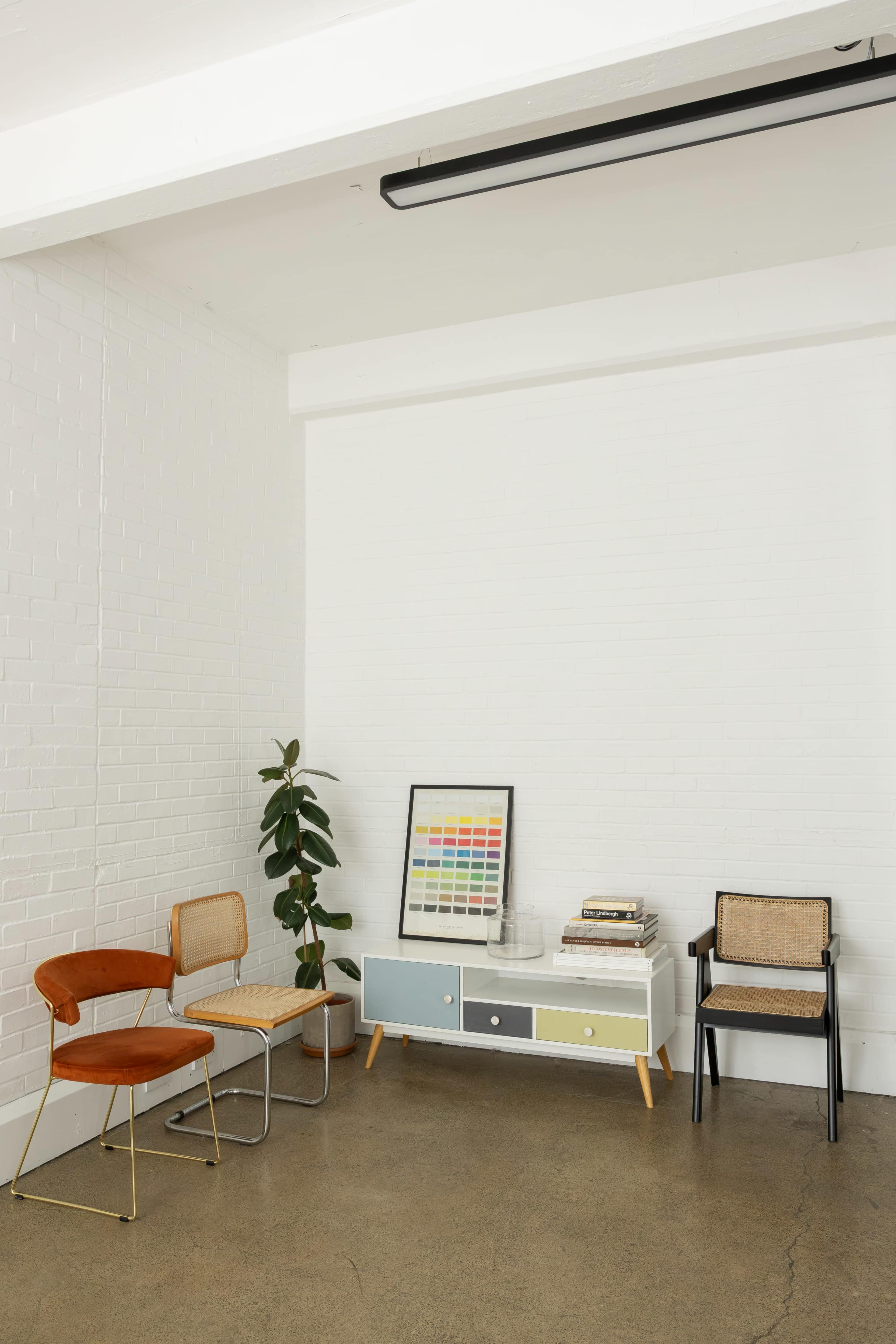The image shows a minimalist interior with two distinct chairs, a low cabinet with colored drawers, a framed color chart, and a potted plant against a white brick wall.