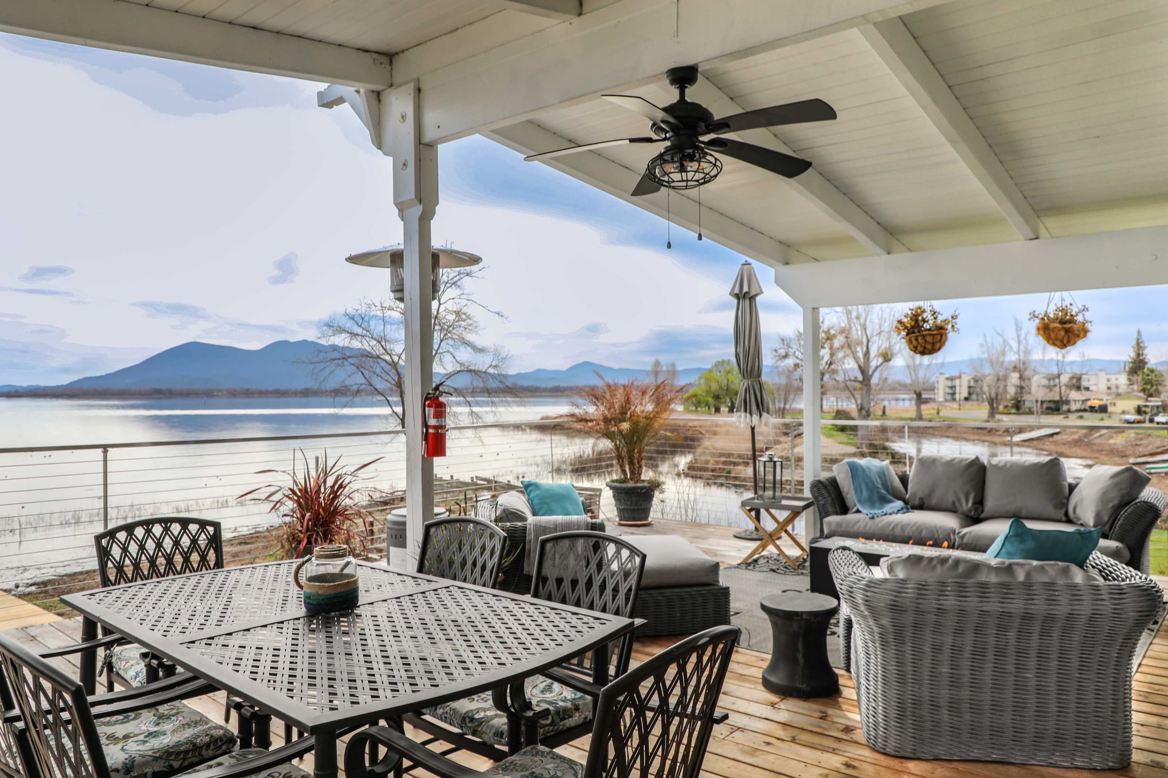 A covered outdoor patio with a dining table, seating area, and a view of a lake and mountains in the background.