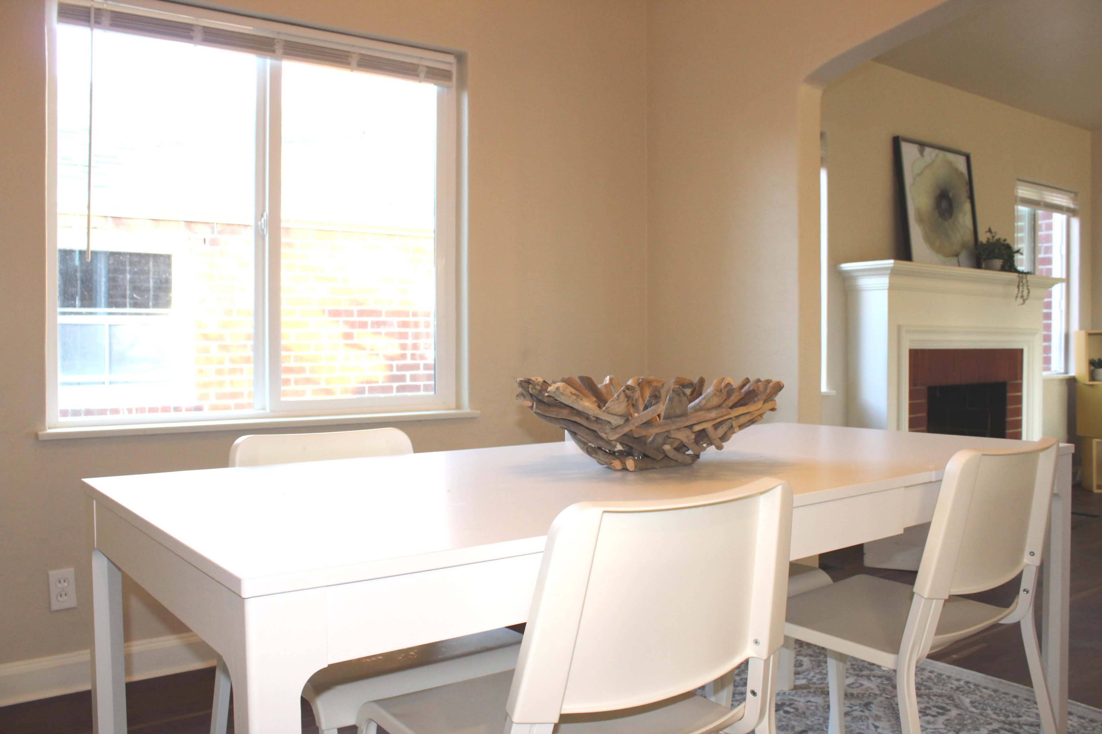 A rectangular white dining table with four white chairs sits under a window, featuring a decorative bowl in the center.