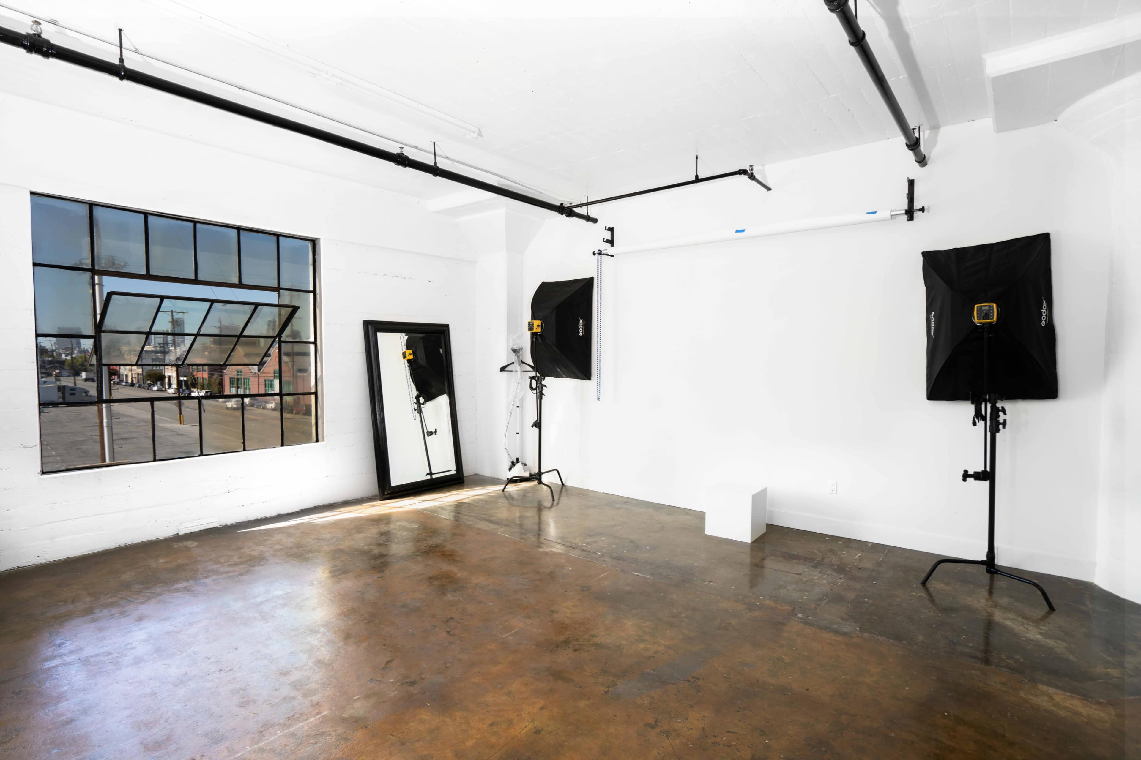 A spacious, empty room with large windows, a mirror, and two lighting setups on stands.