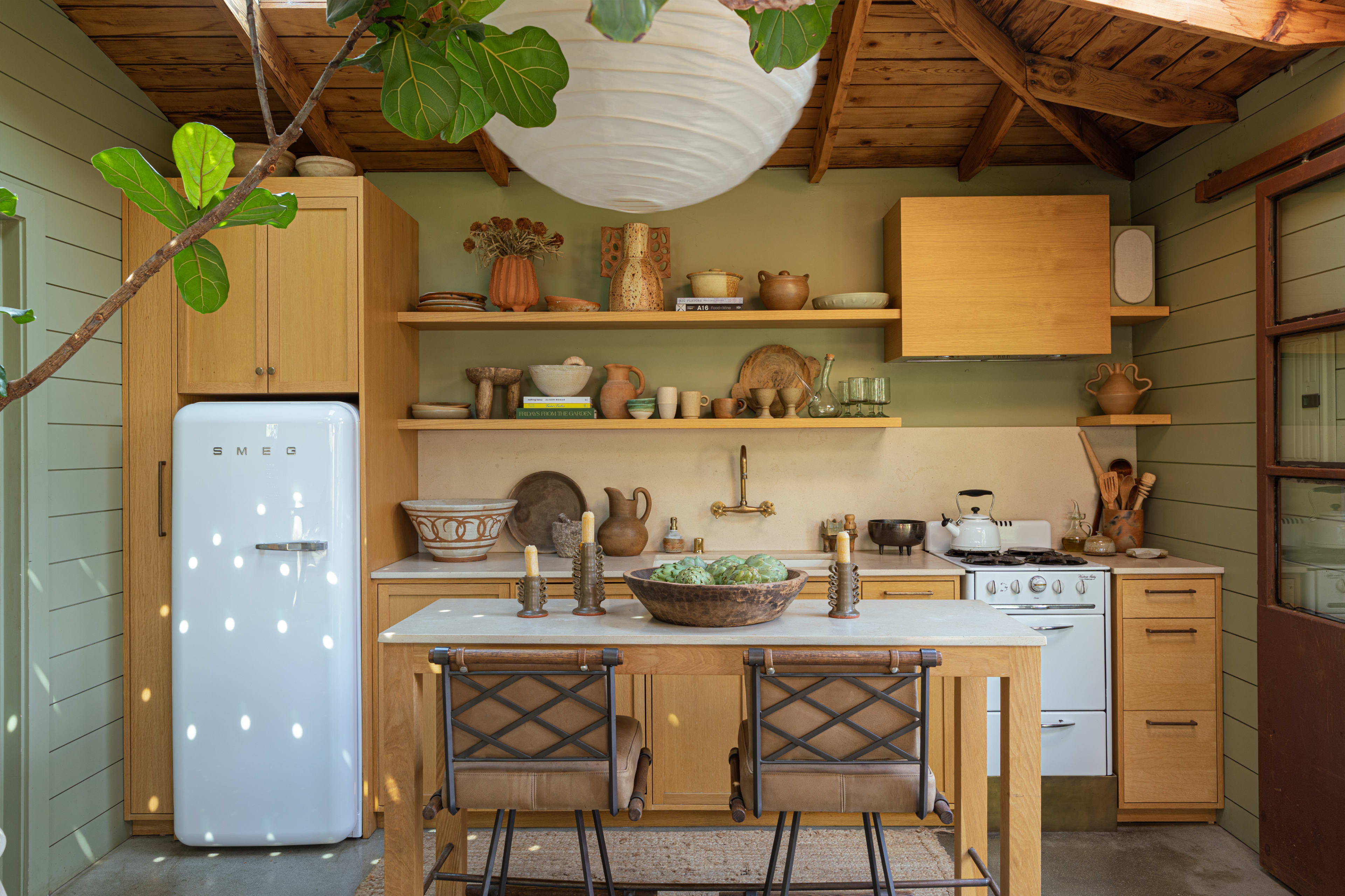 The image depicts a rustic kitchen with wooden shelves displaying various pottery items, a white refrigerator, and a wooden table with two chairs, all under a wooden-beamed ceiling.