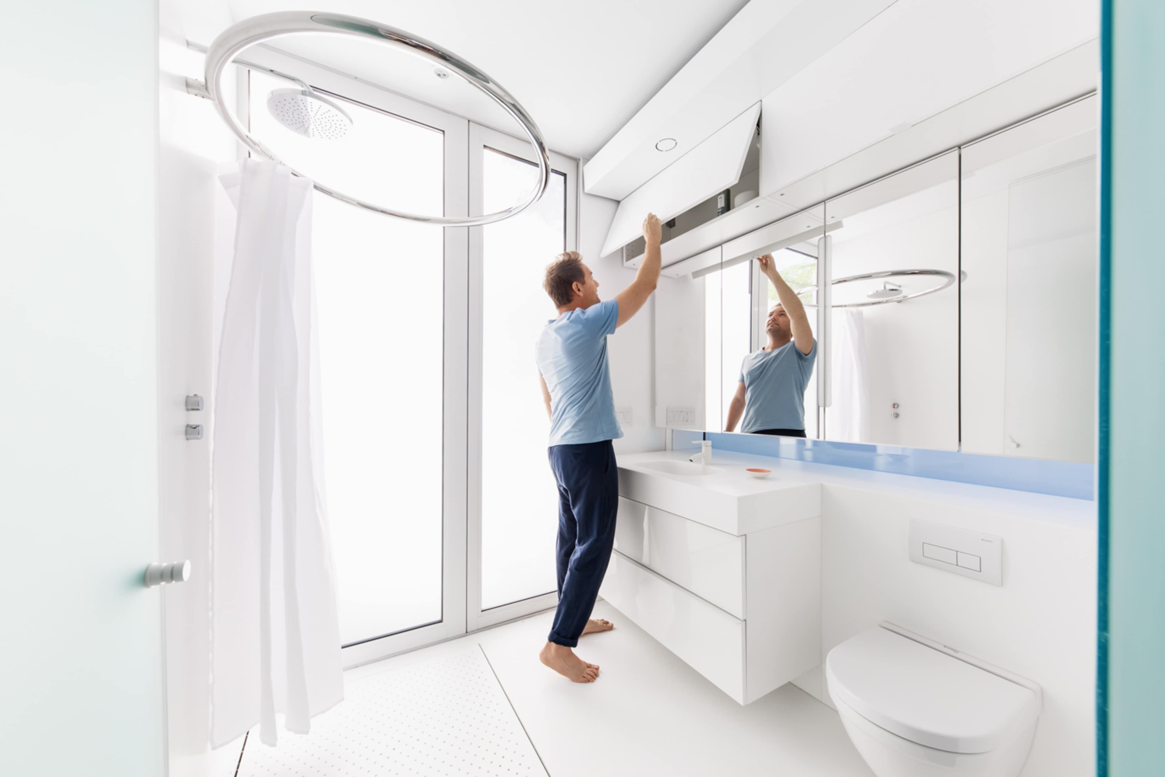 A man in a light blue shirt adjusts a mirror above a modern bathroom vanity with a minimalist design.