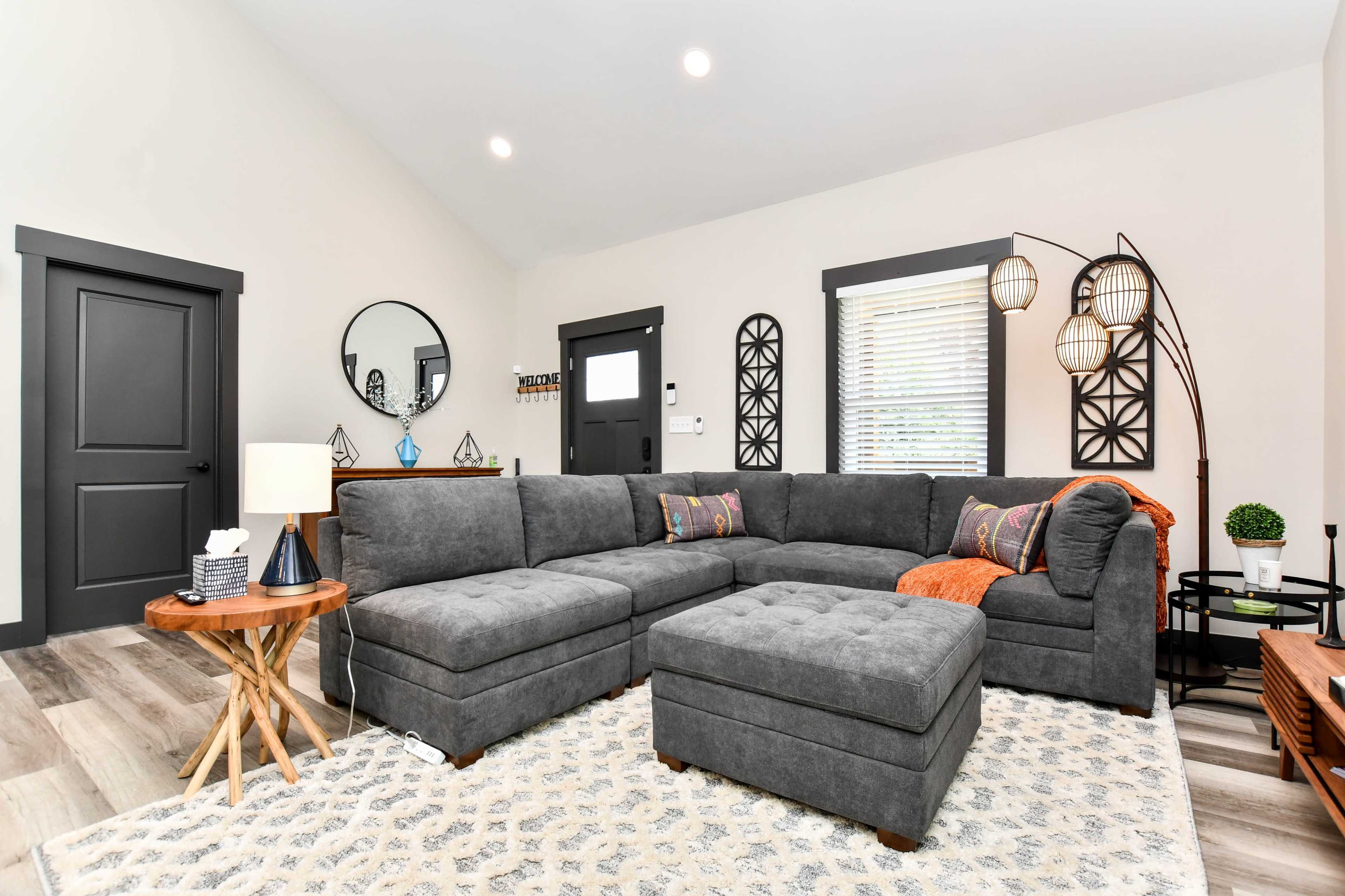 The living room features a gray sectional sofa with an orange throw blanket, a round mirror on the wall, and a wooden coffee table beside a decorative lamp.
