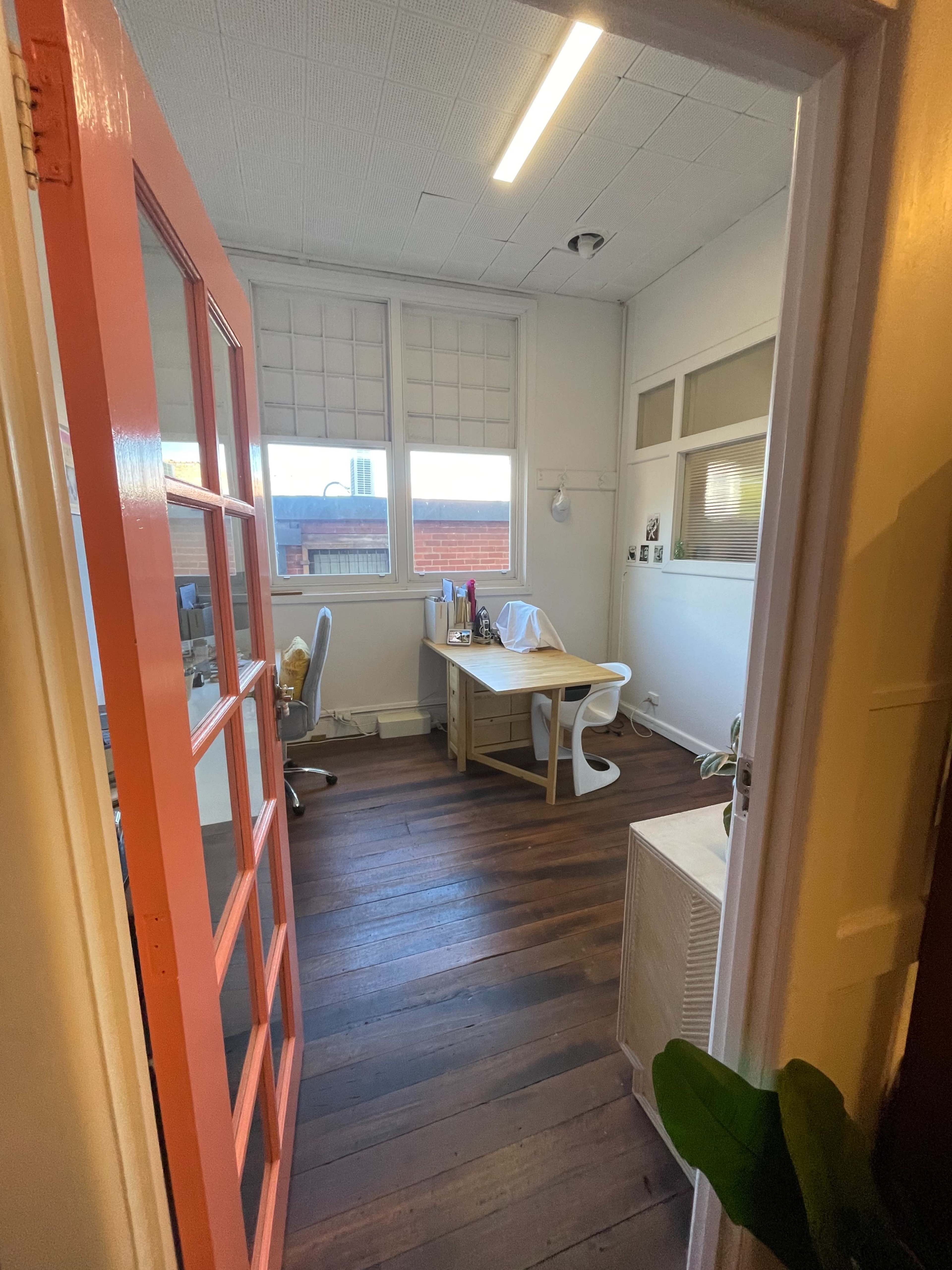 The image shows a narrow office space with wooden floors, a desk and chair, and large windows with blinds.