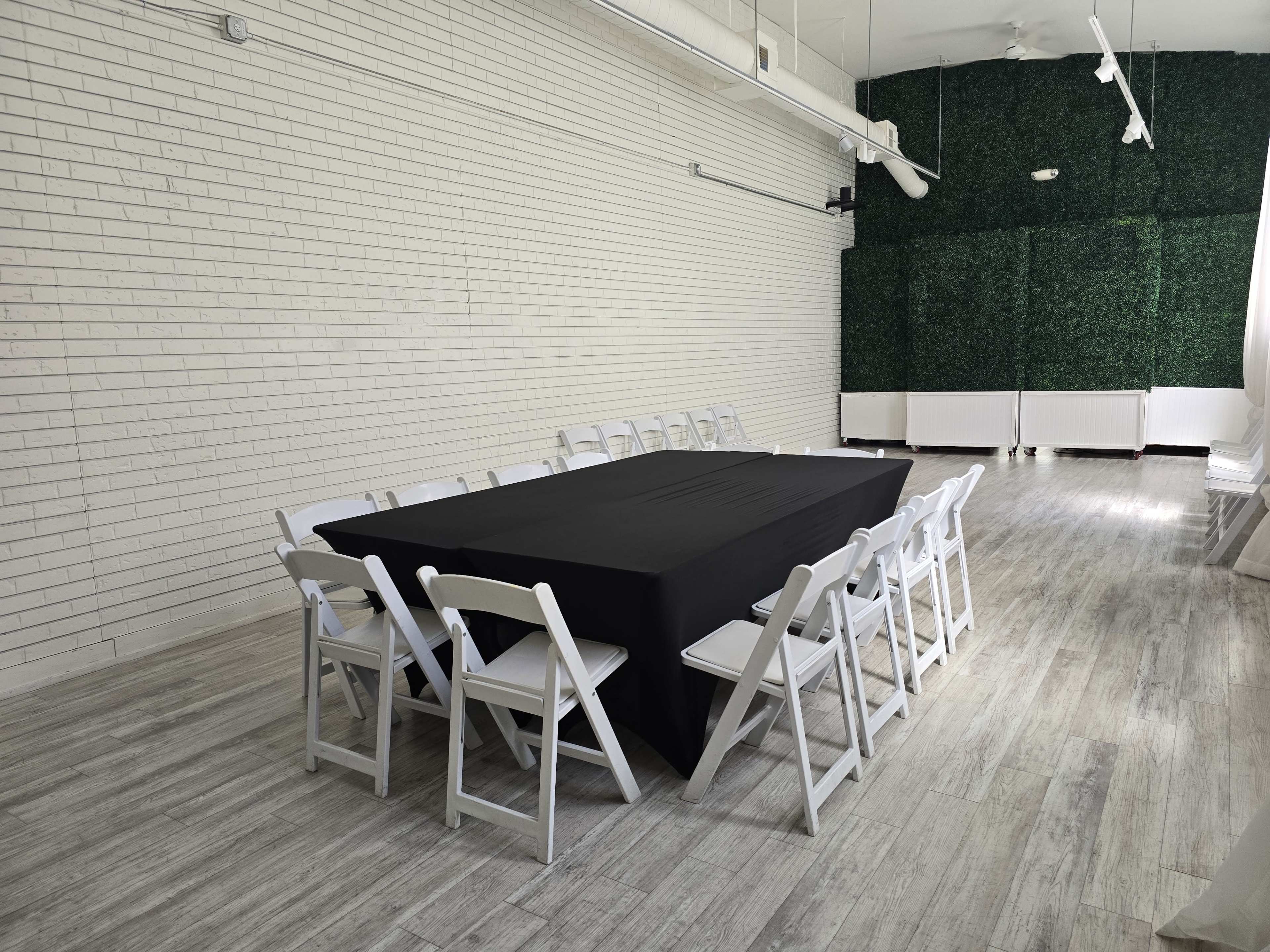 A long black table surrounded by white folding chairs is set up in a bright, minimalist room with white brick walls and a green foliage backdrop.