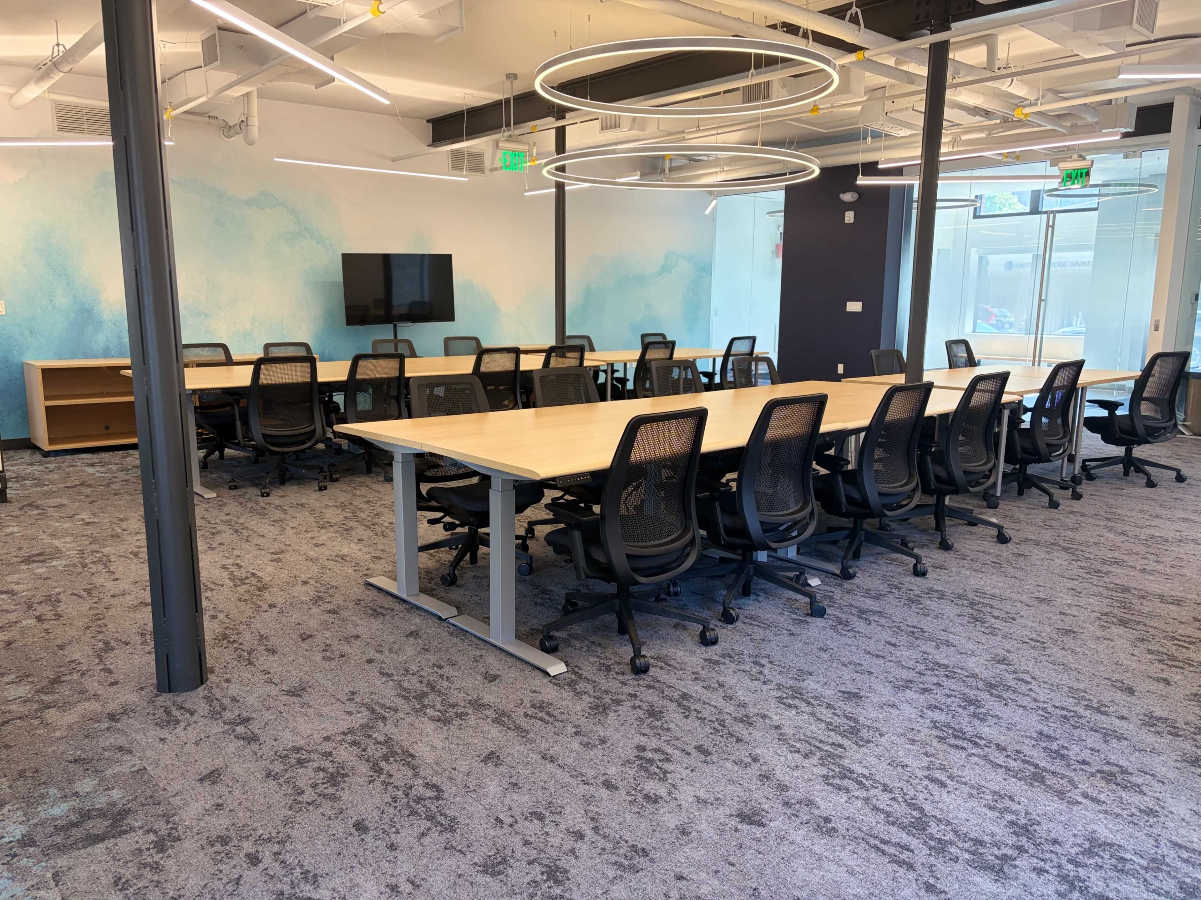 The image shows a modern conference room with a long table surrounded by black chairs, a large screen on the wall, and a light blue mural in the background.