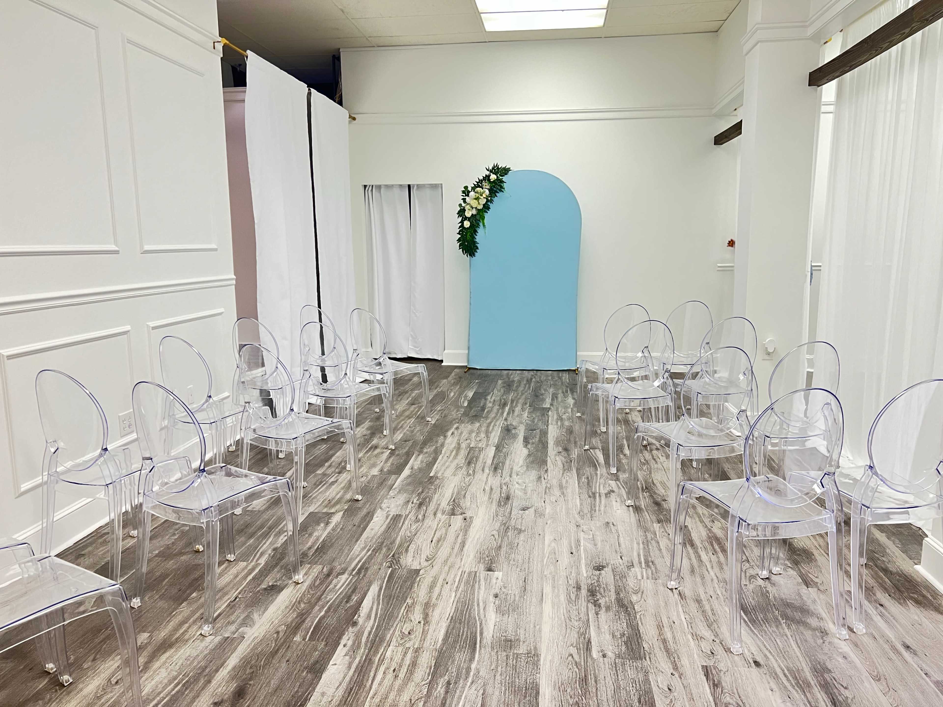 A minimalist indoor space arranged with transparent chairs on both sides of a central path leading to a blue arch adorned with floral elements.