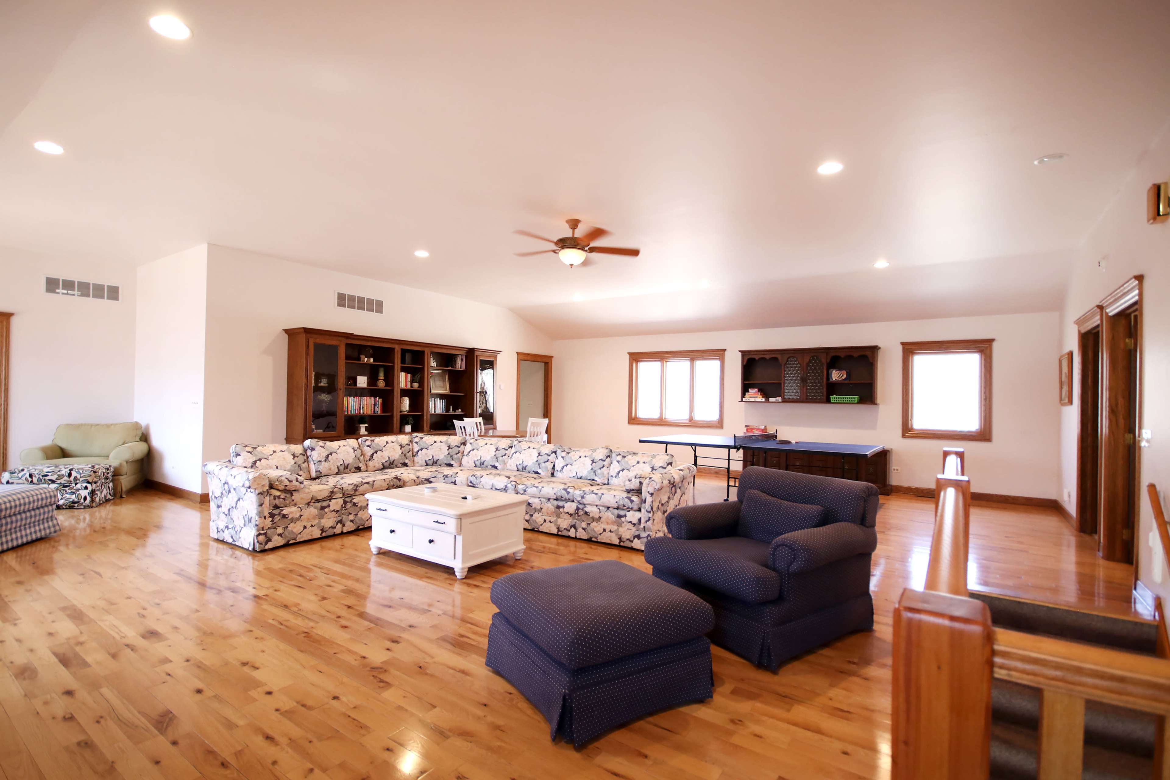 The spacious living room features a floral-patterned sectional sofa, a wooden bookcase, and a ceiling fan, all set against polished hardwood floors.