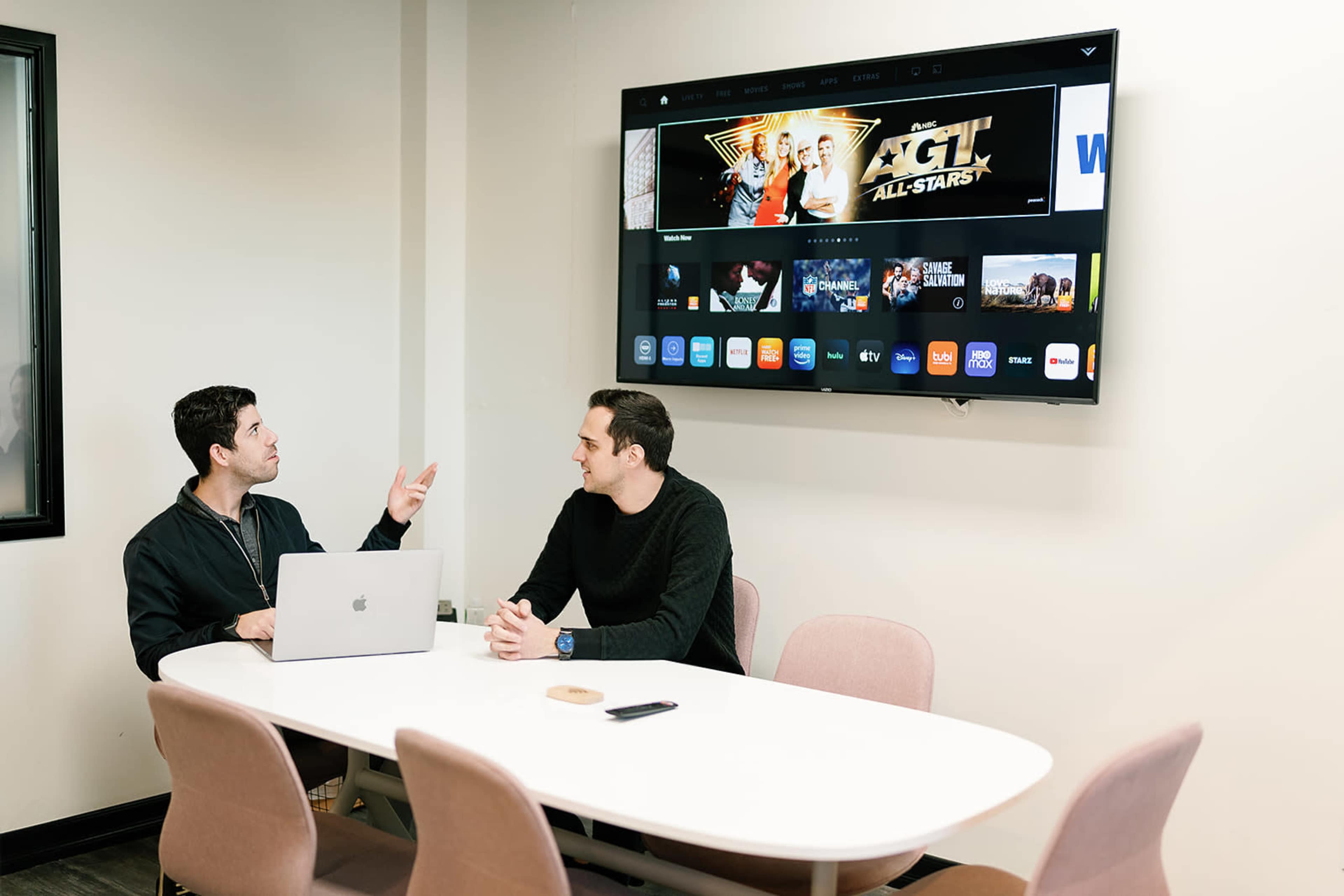 Two men are seated at a table in a meeting room, discussing topics while a large screen displays various media options.