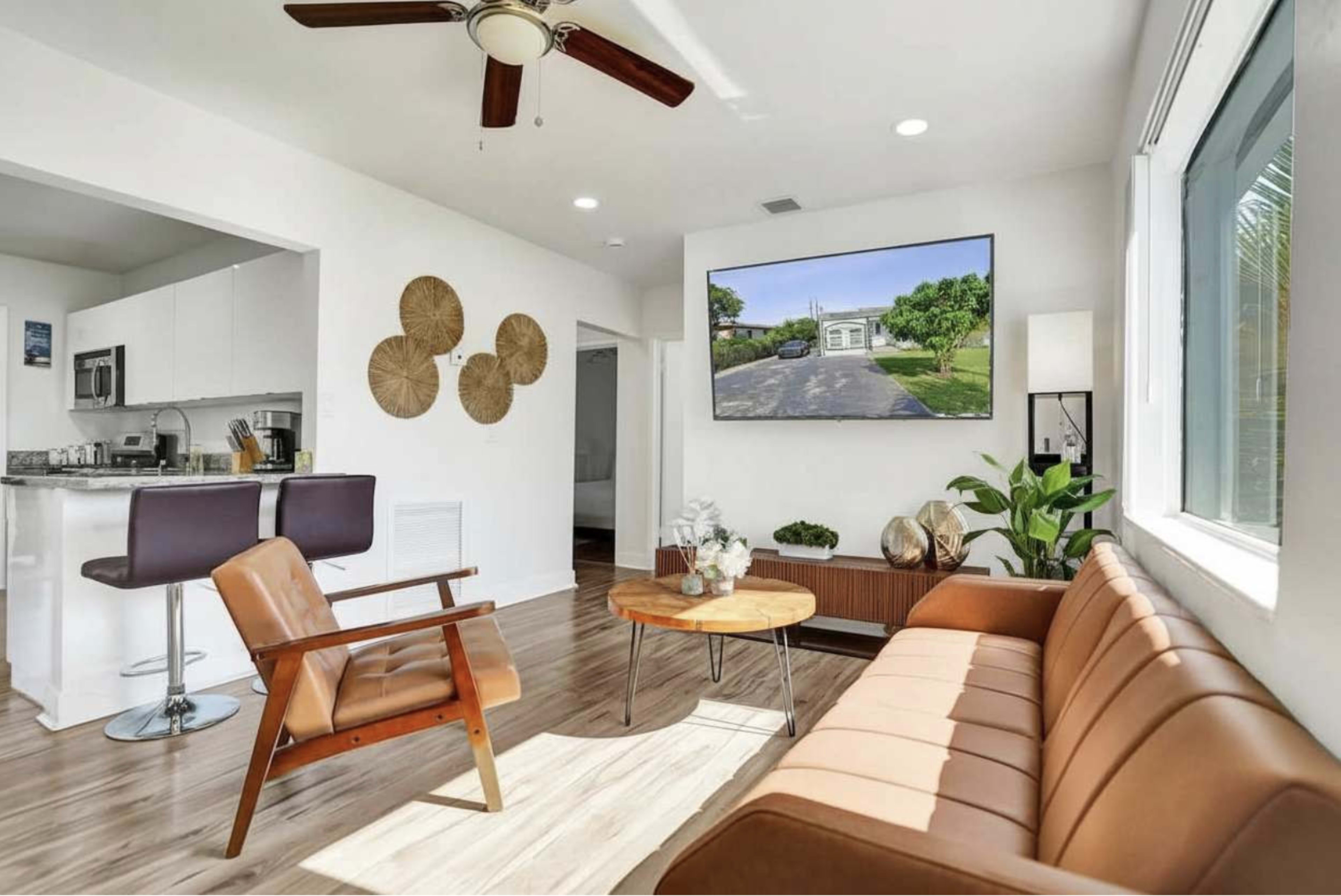 The image features a modern living room with a brown leather sofa, a round wooden coffee table, and a large wall-mounted television displaying an outdoor scene.