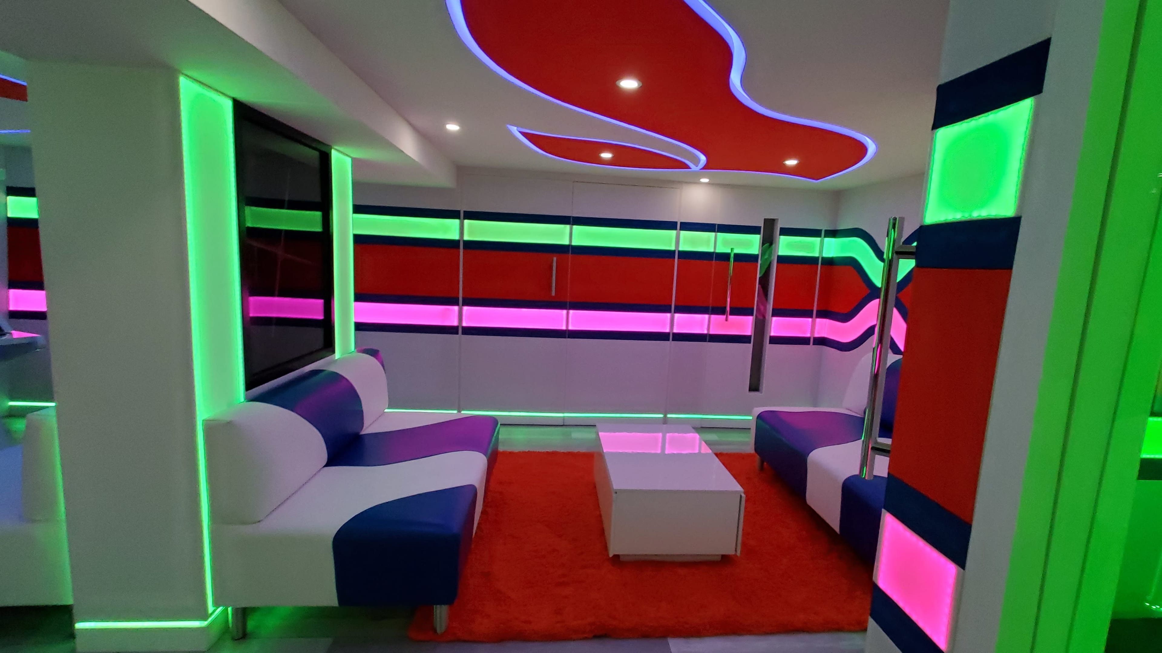 A modern living room with vibrant, multicolored wall designs and furniture featuring white and colored couches, a glass coffee table, and illuminated accents.