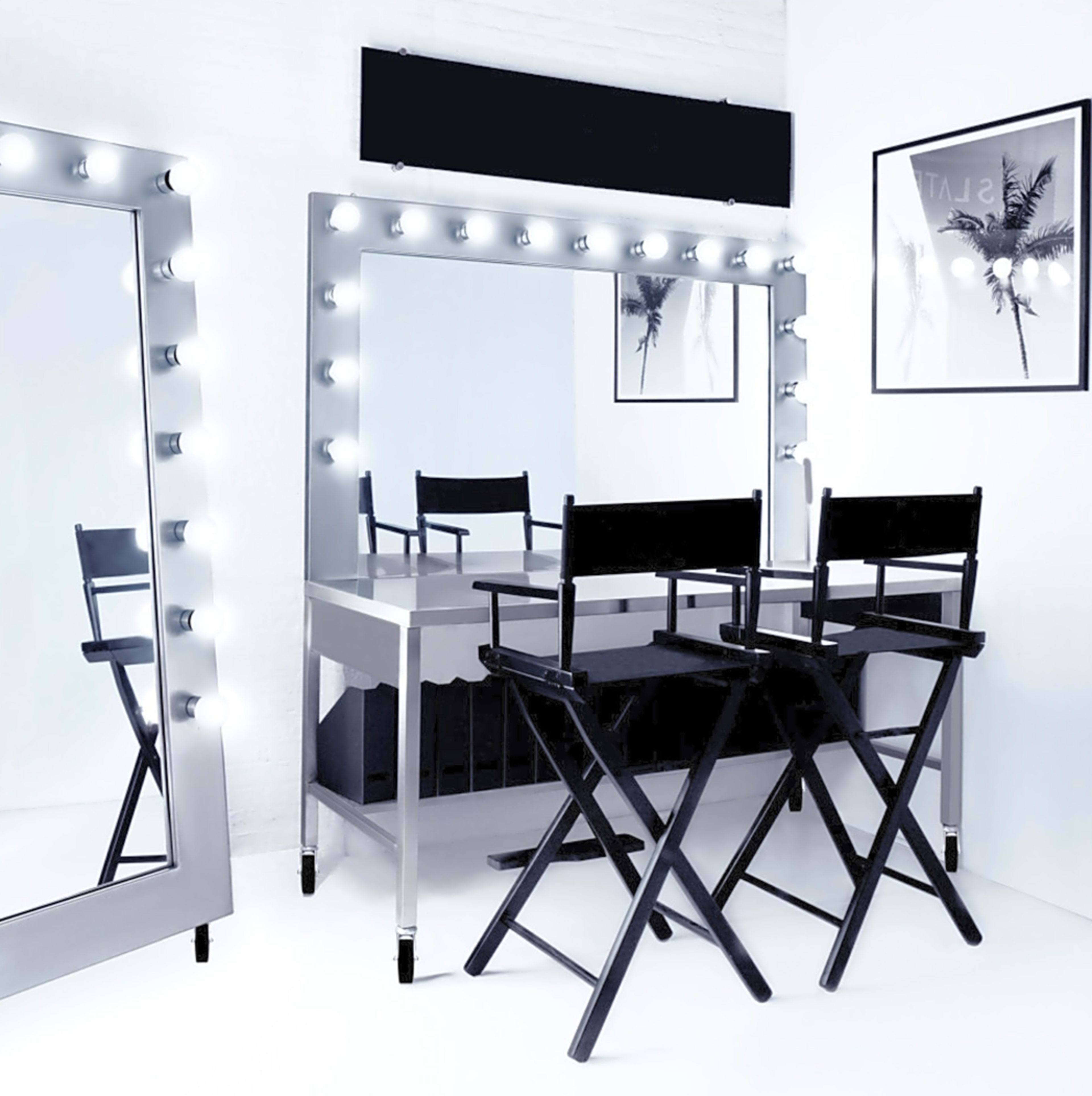 A well-lit makeup station with a large mirror framed by bulbs, accompanied by two black director's chairs and framed photos on the walls.