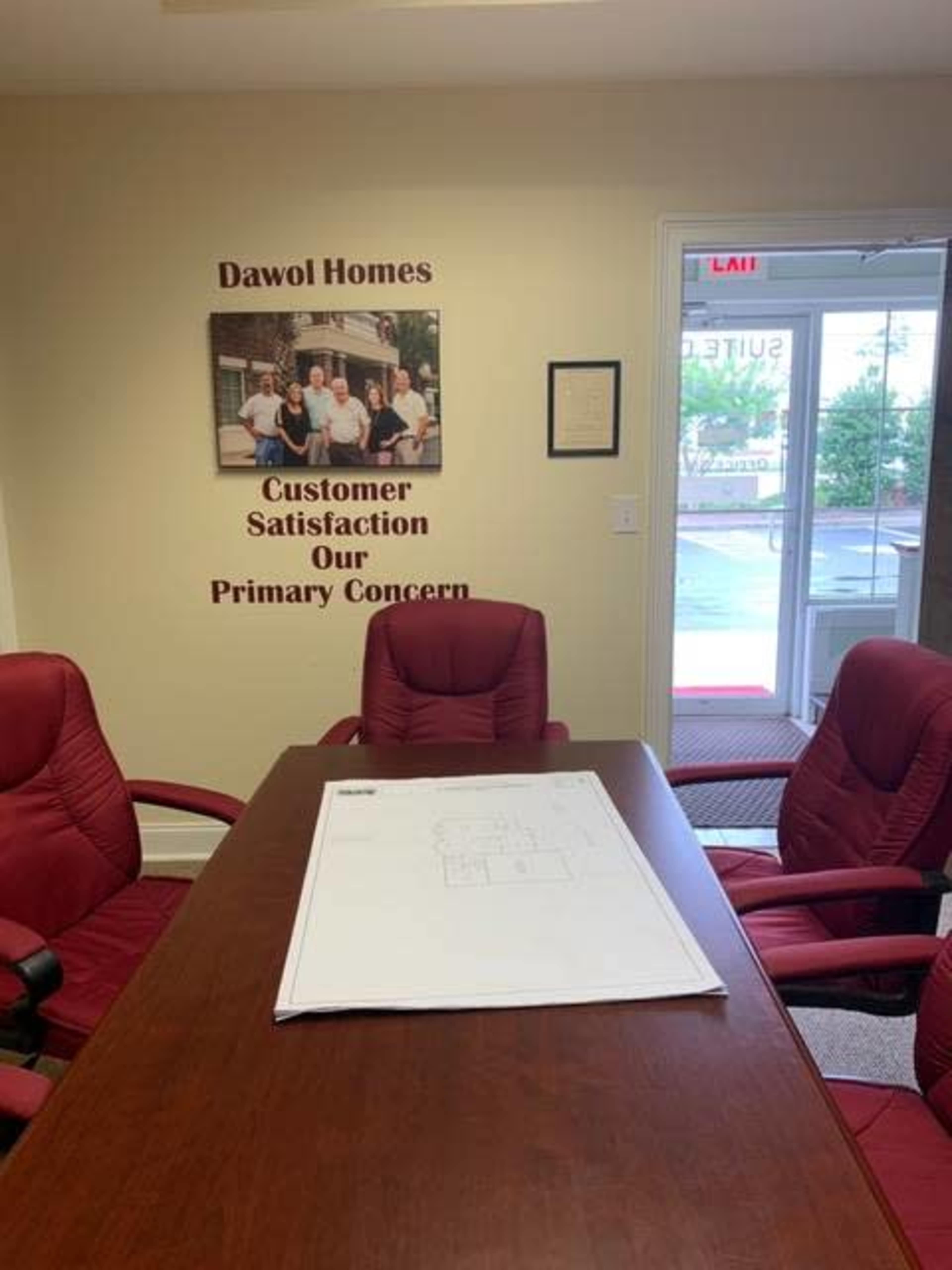 A conference room with a large table, red chairs, a wall displaying "Dawol Homes" and a customer service motto, and a blueprint on the table.