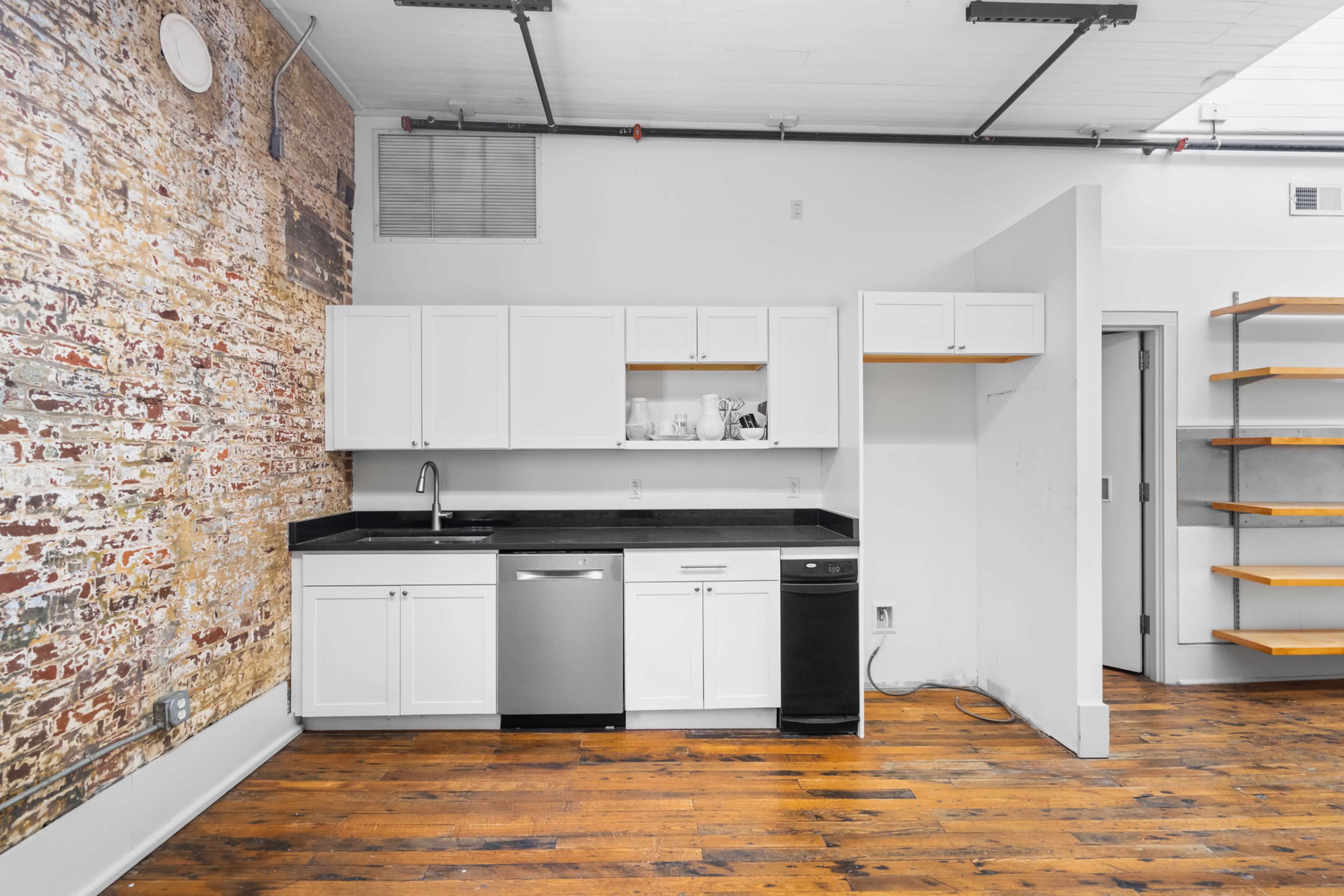 The image shows a minimalist kitchen with white cabinets, a black countertop, stainless steel appliances, and an exposed brick wall in an open space.