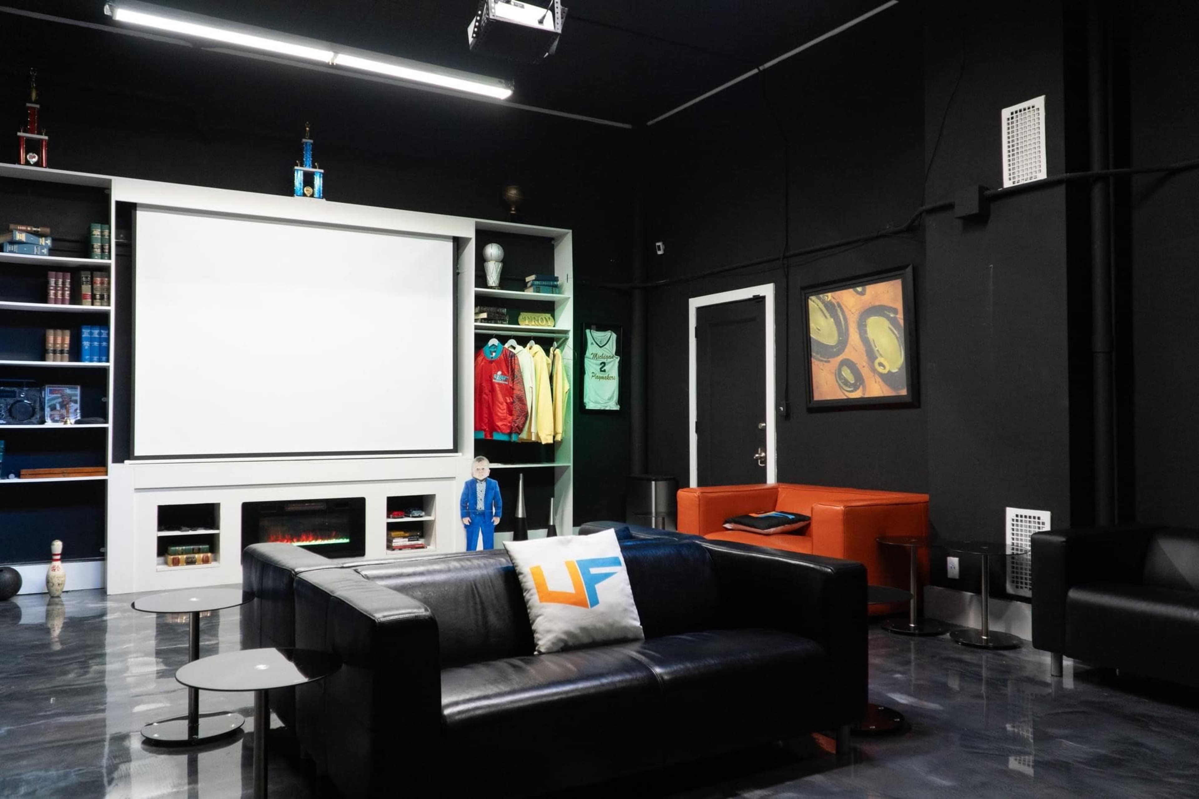 A modern lounge featuring a black leather sofa, a large white screen on the wall, and shelves filled with books and colorful clothing items.