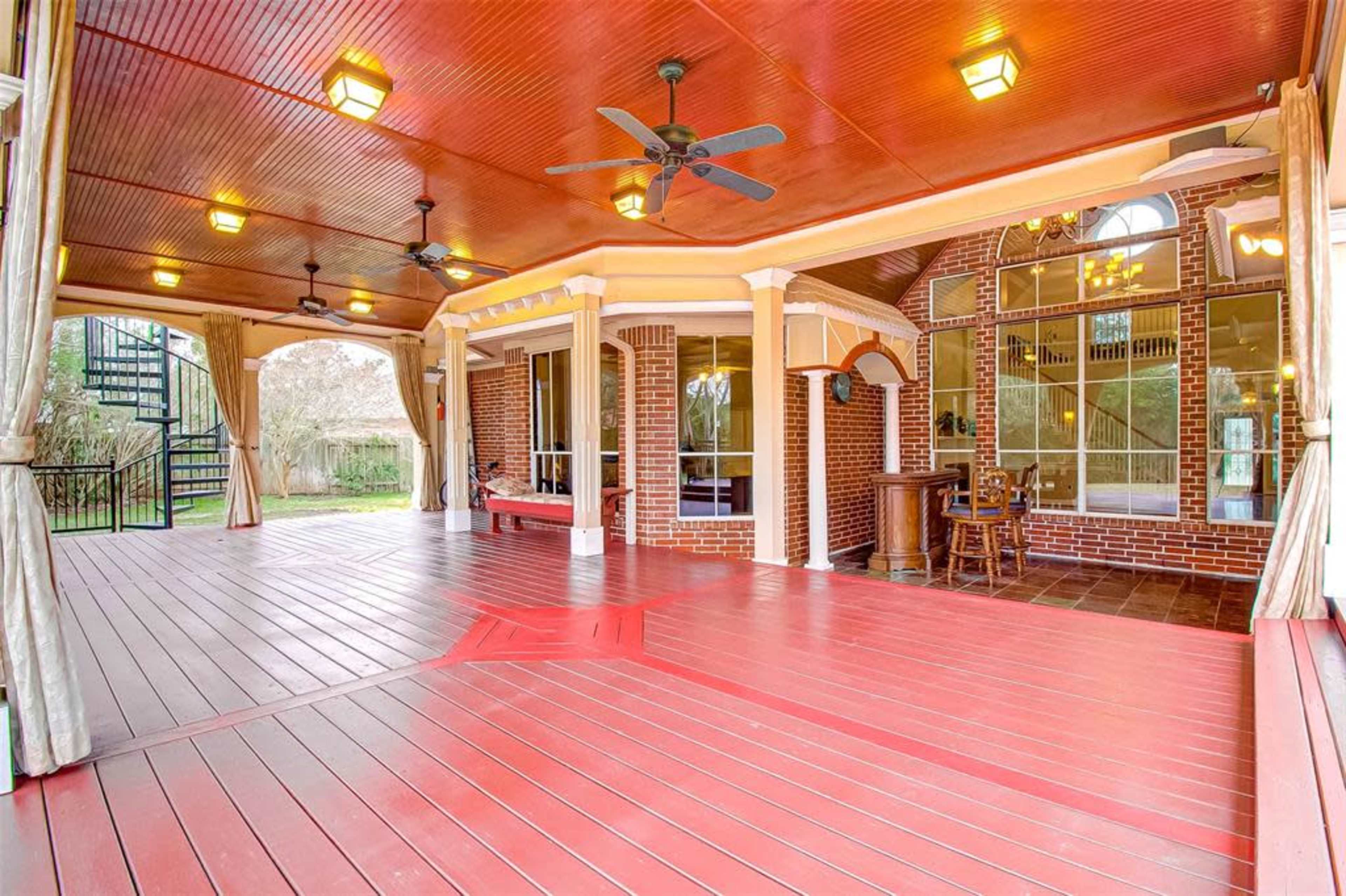 A spacious covered patio with wooden flooring, ceiling fans, and brick columns, featuring a spiral staircase leading to an upper level.