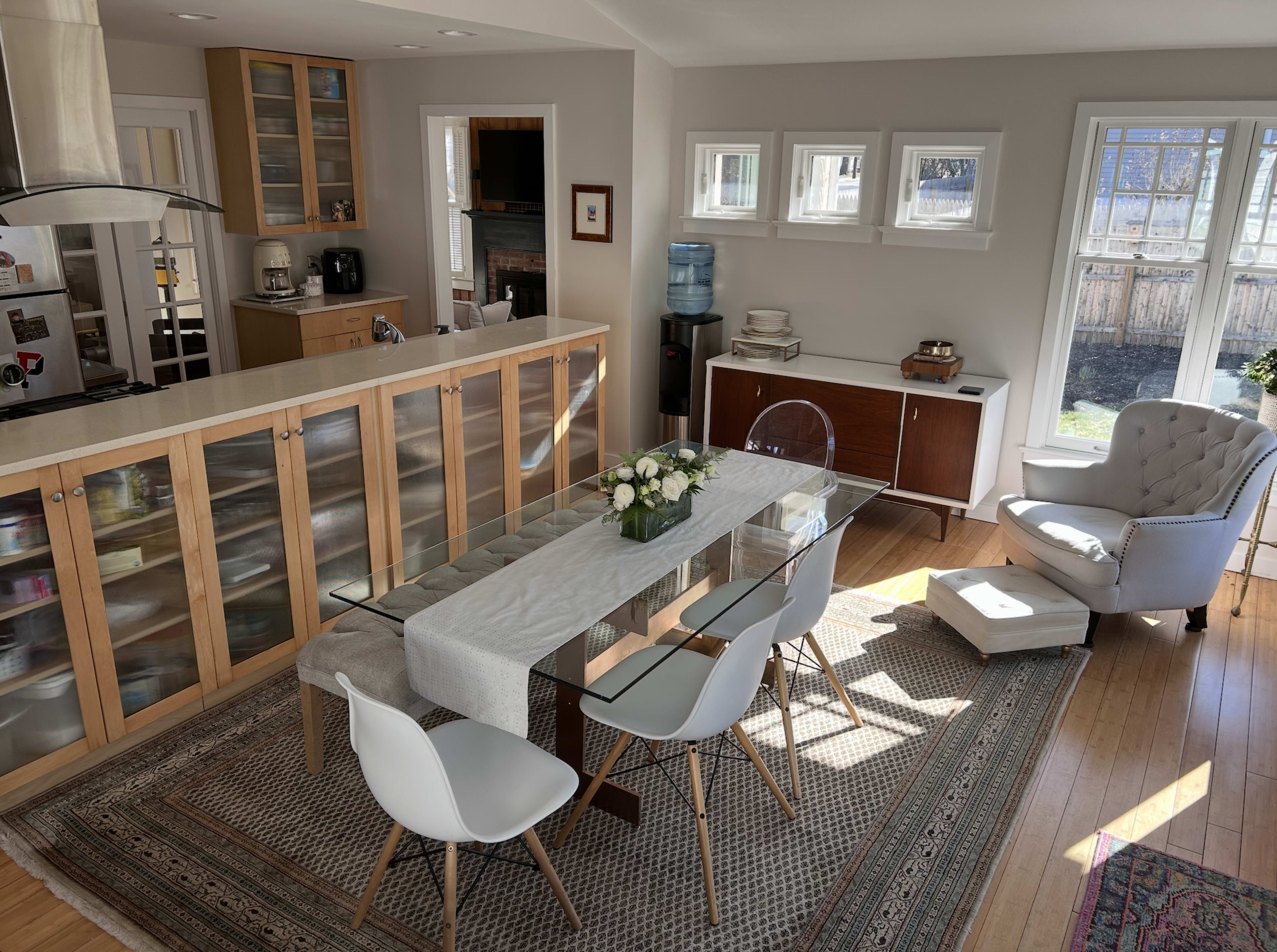 The image shows a bright dining area featuring a glass table with white chairs, a floral centerpiece, and a cozy seating arrangement near windows.