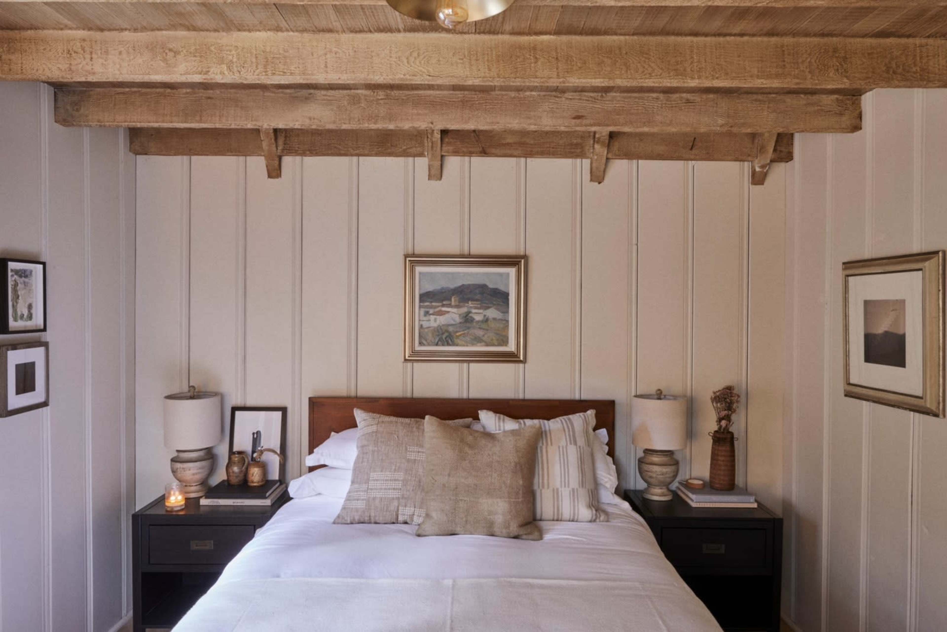 A cozy bedroom featuring a wooden ceiling with exposed beams, a neatly made bed, and bedside tables with lamps and framed artwork on the walls.