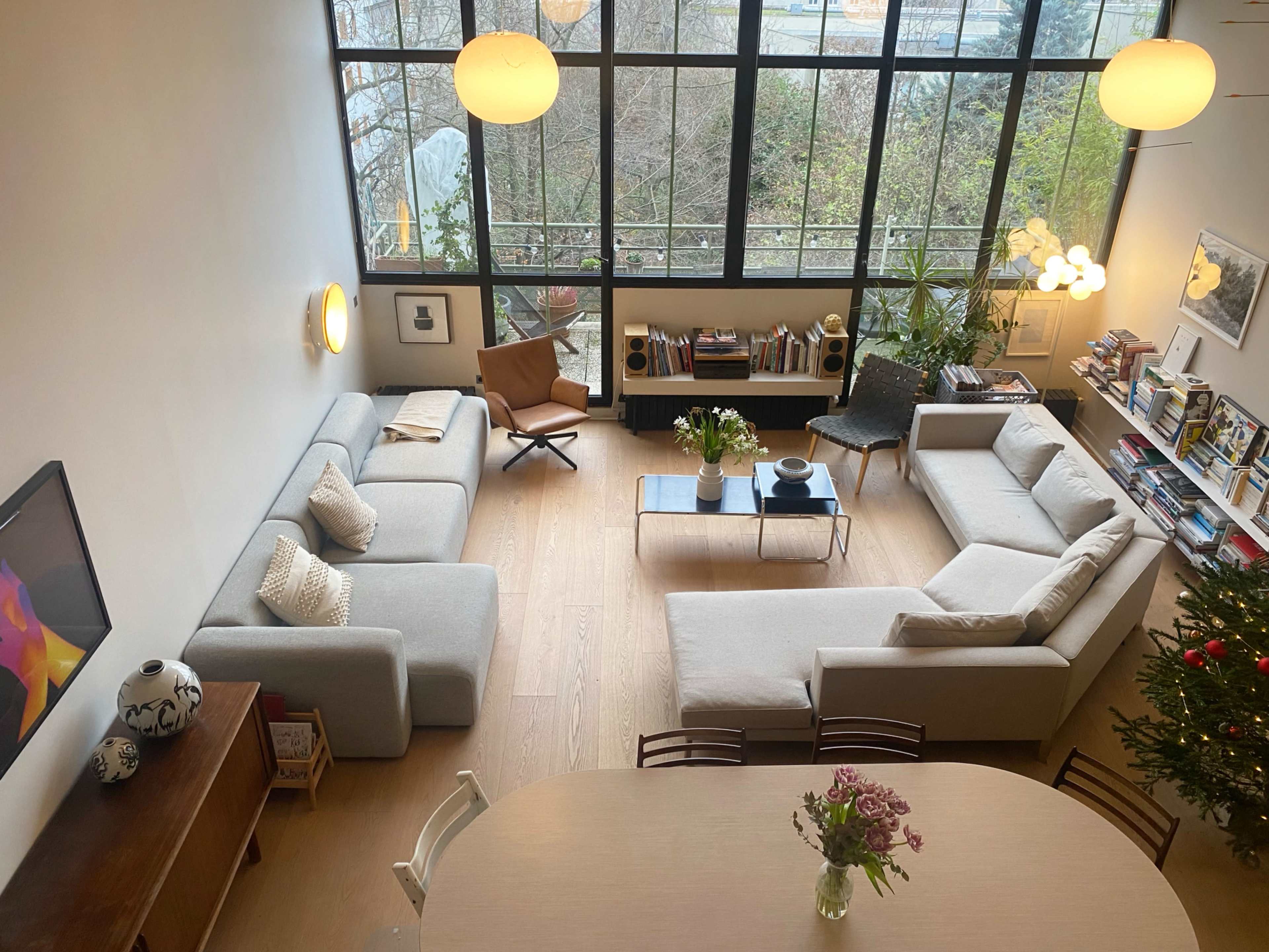 The image shows a modern living room with large windows, featuring a sectional sofa, a coffee table, and bookshelves, along with a dining area and indoor plants.