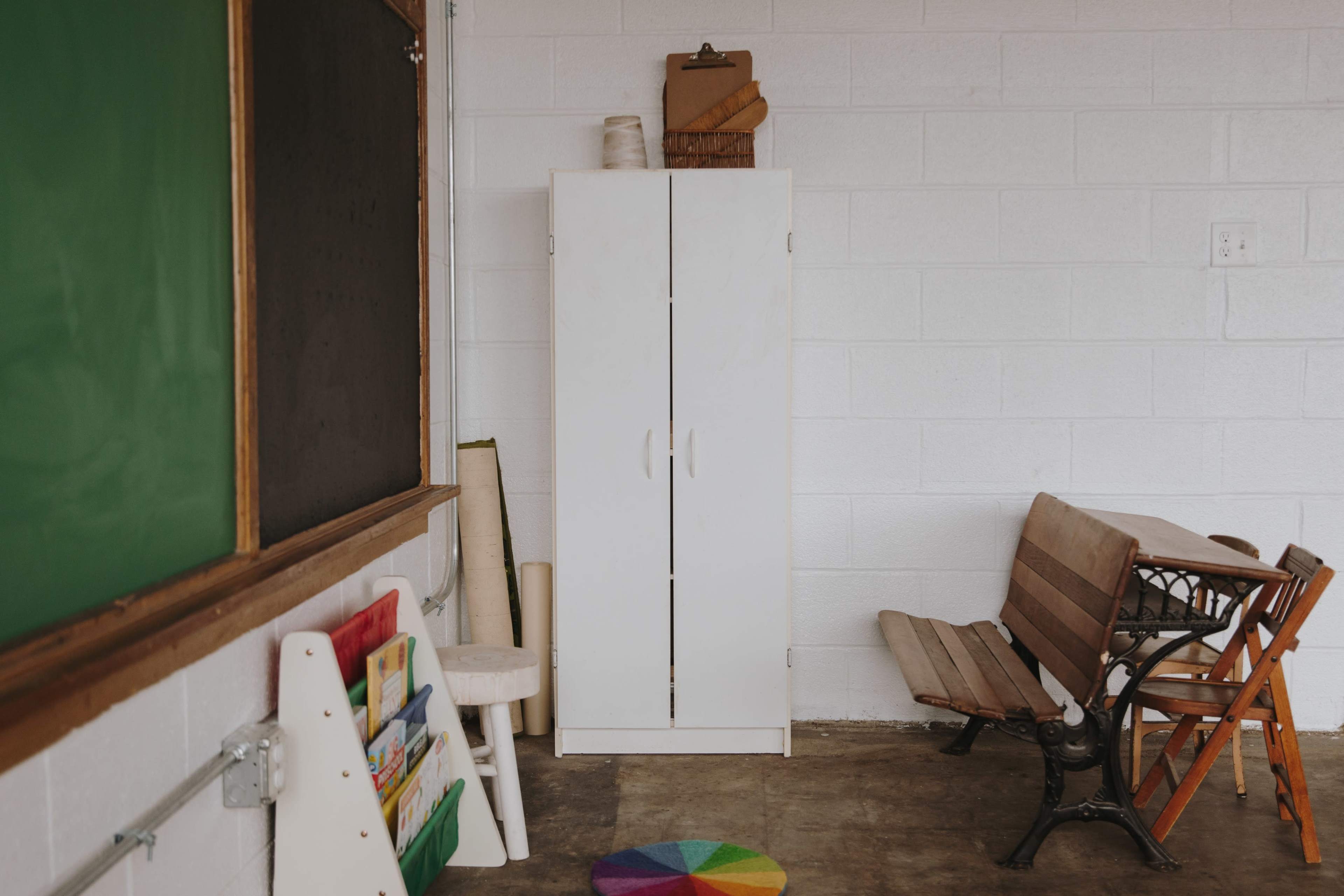 The image shows a white storage cabinet in a classroom corner next to a green chalkboard and wooden benches.