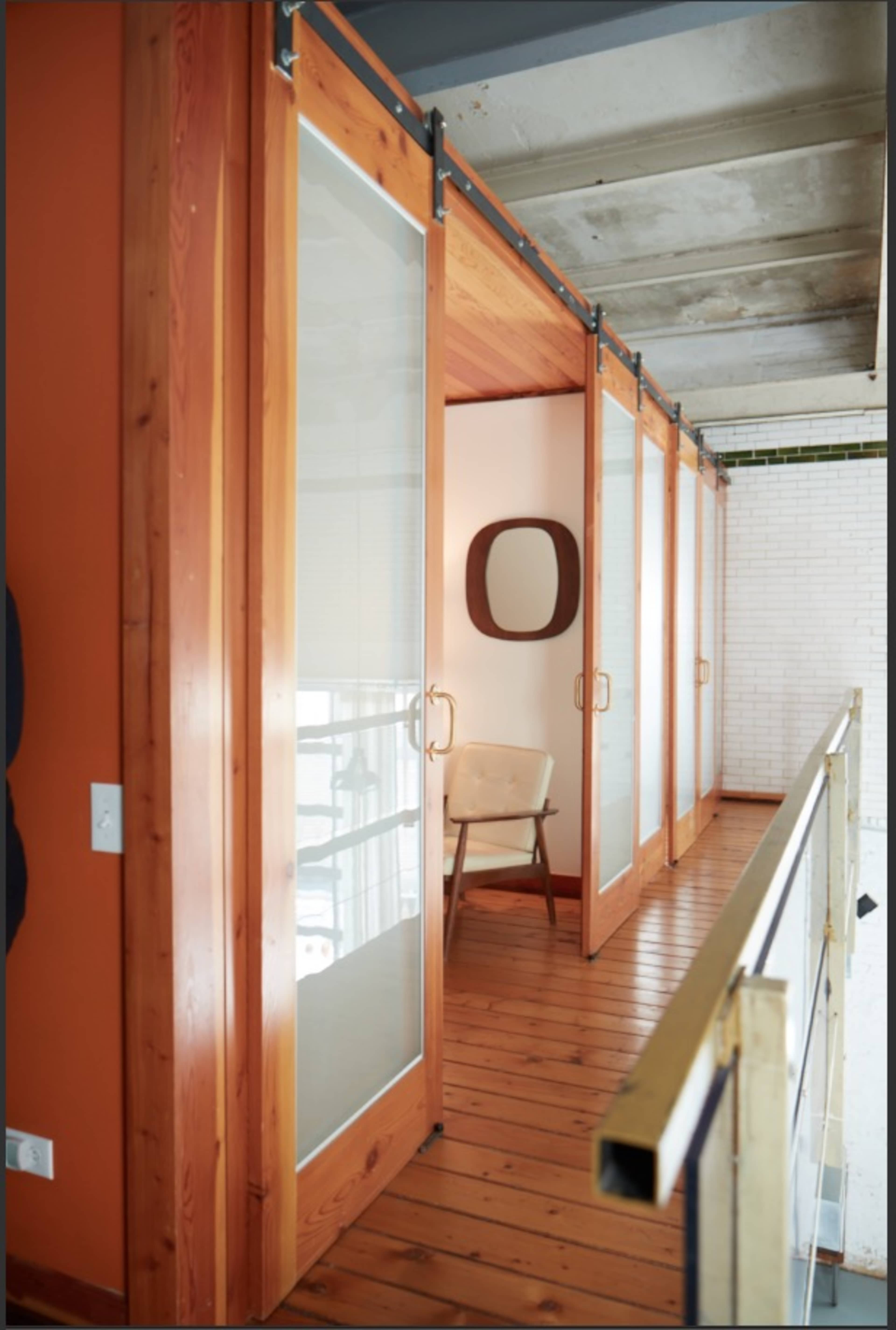 The image shows a hallway with wooden sliding doors and a simple chair, characterized by exposed beams and a contrasting orange wall.