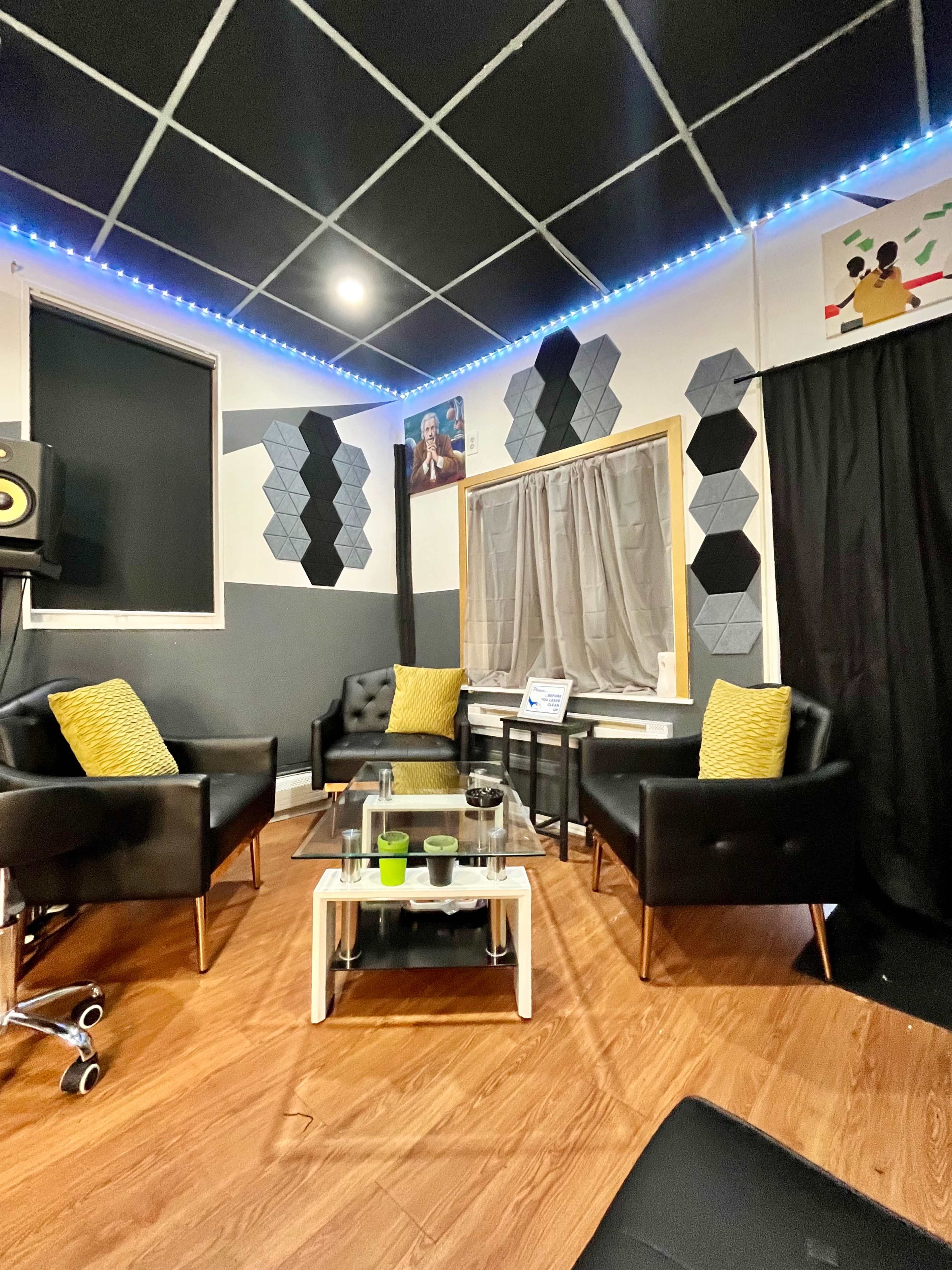The image shows a cozy lounge area with two black armchairs, a glass coffee table, and decorative hexagonal wall panels, illuminated by blue LED lights.