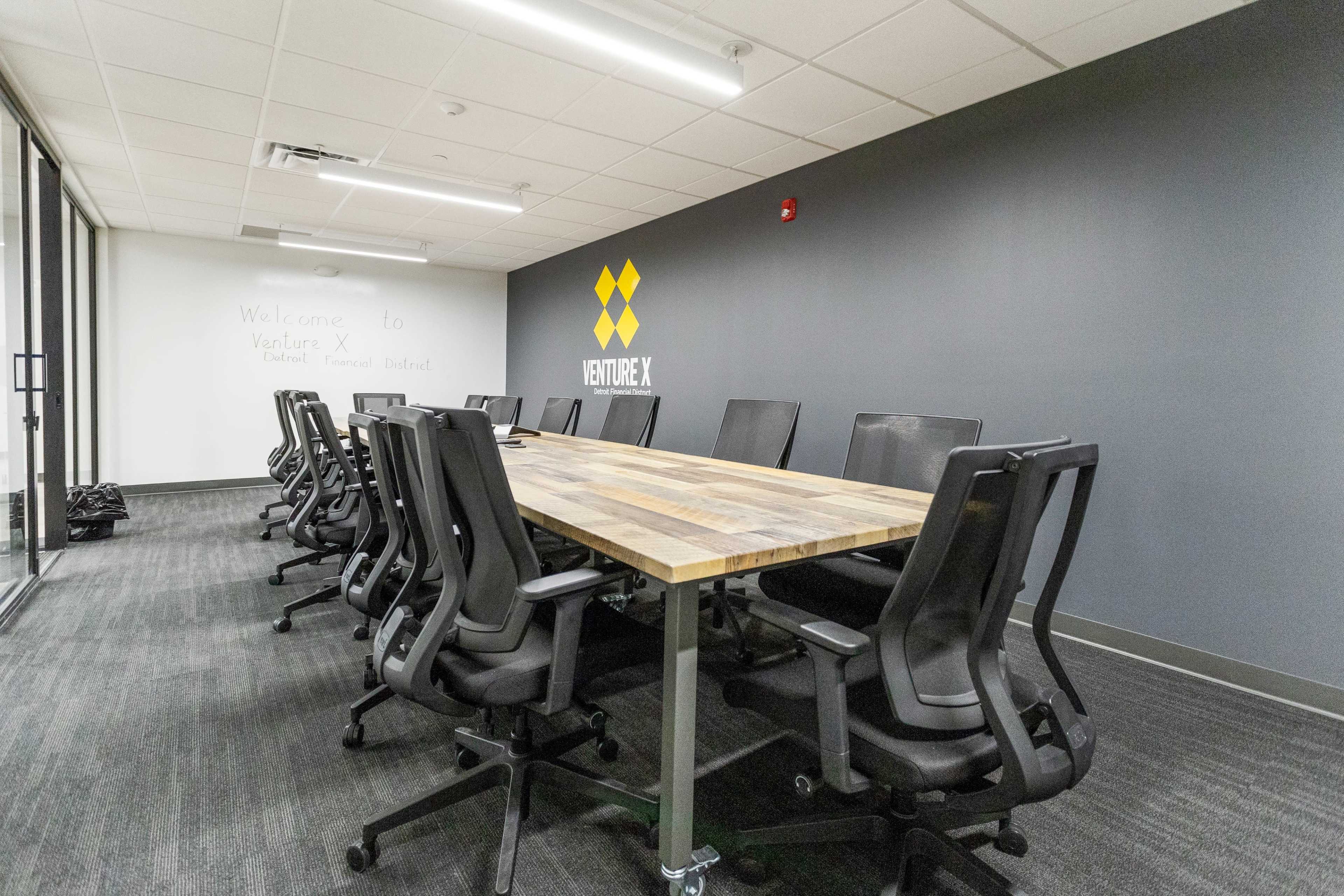 A modern conference room features a long wooden table surrounded by black ergonomic chairs, with a large logo on the gray wall.