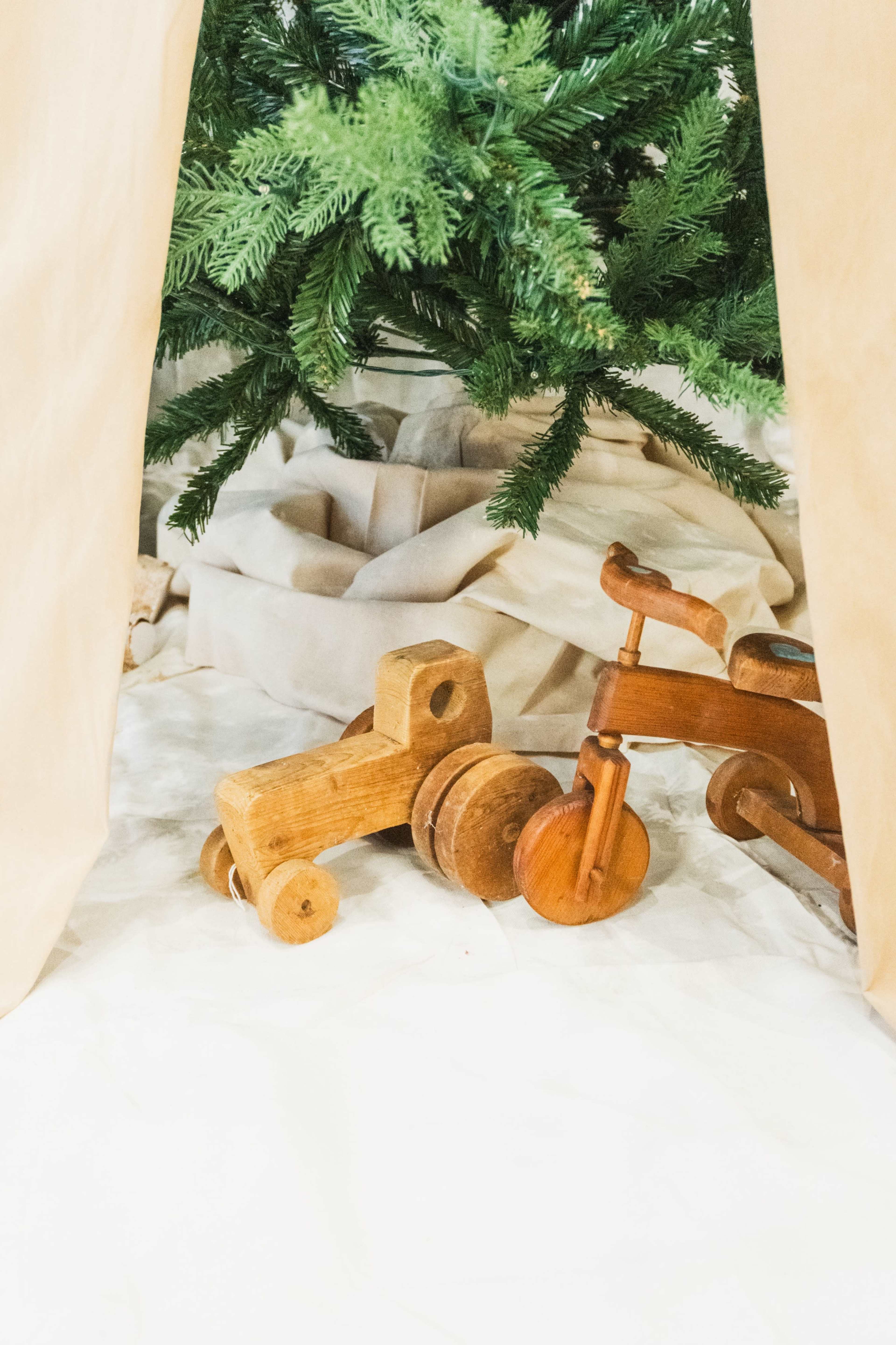 The image features two wooden toys, a push-along truck and a tricycle, positioned beneath a decorated Christmas tree.