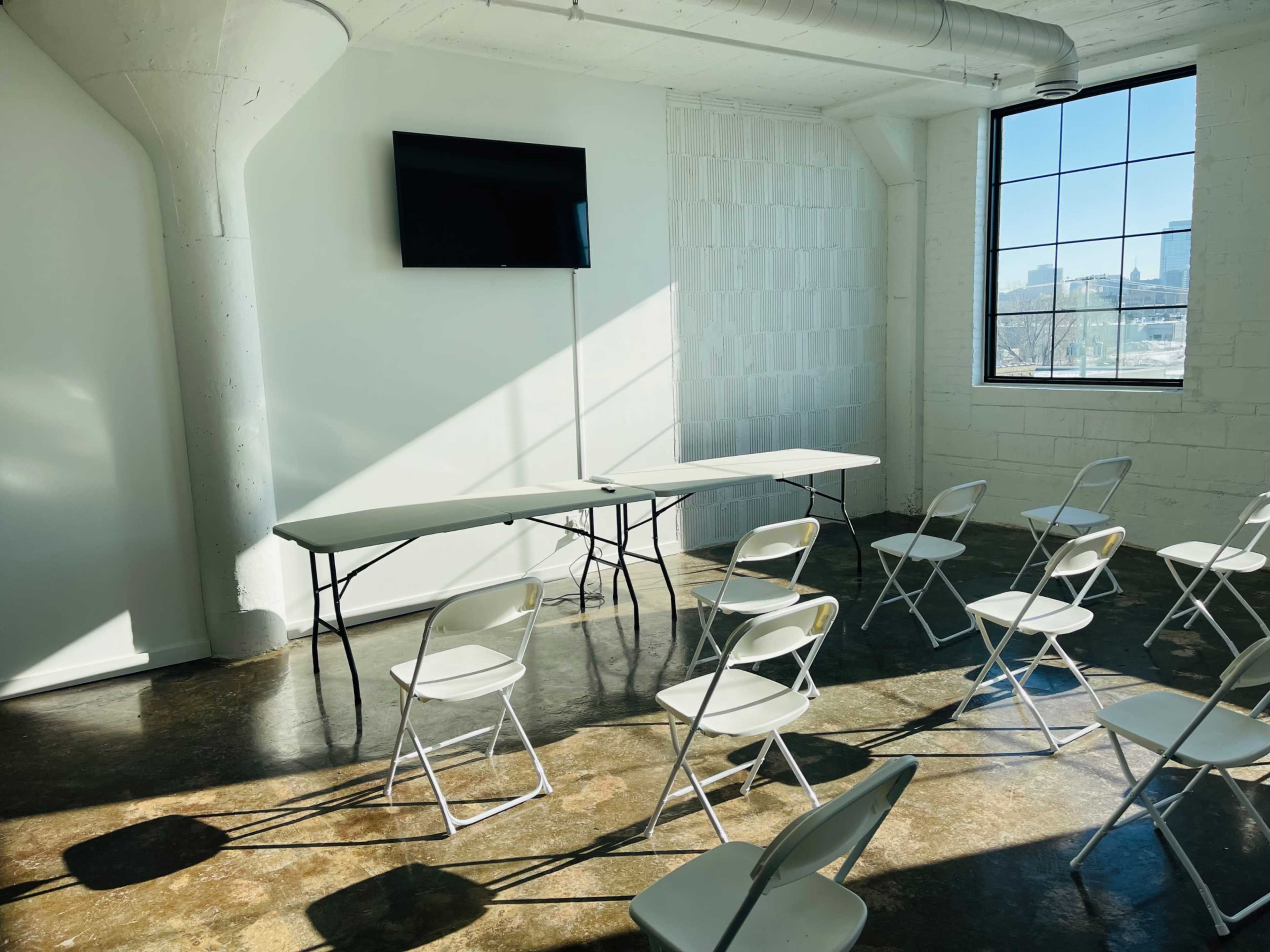 A well-lit meeting room features a table with chairs arranged in front of a wall-mounted television and a large window.