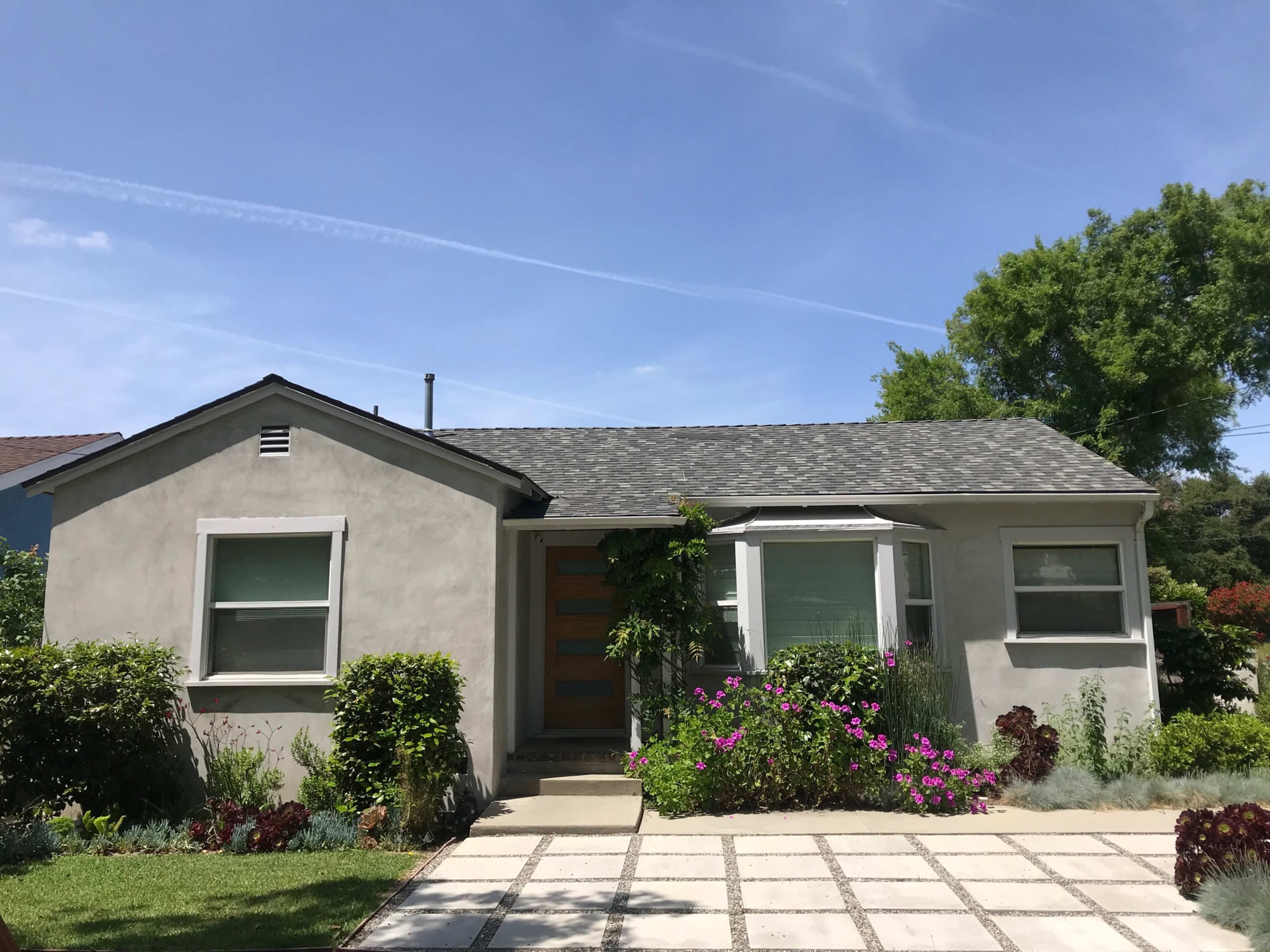 A single-story home with a gray exterior features a front porch, a landscaped garden with colorful flowers, and a stone pathway leading to the entrance.