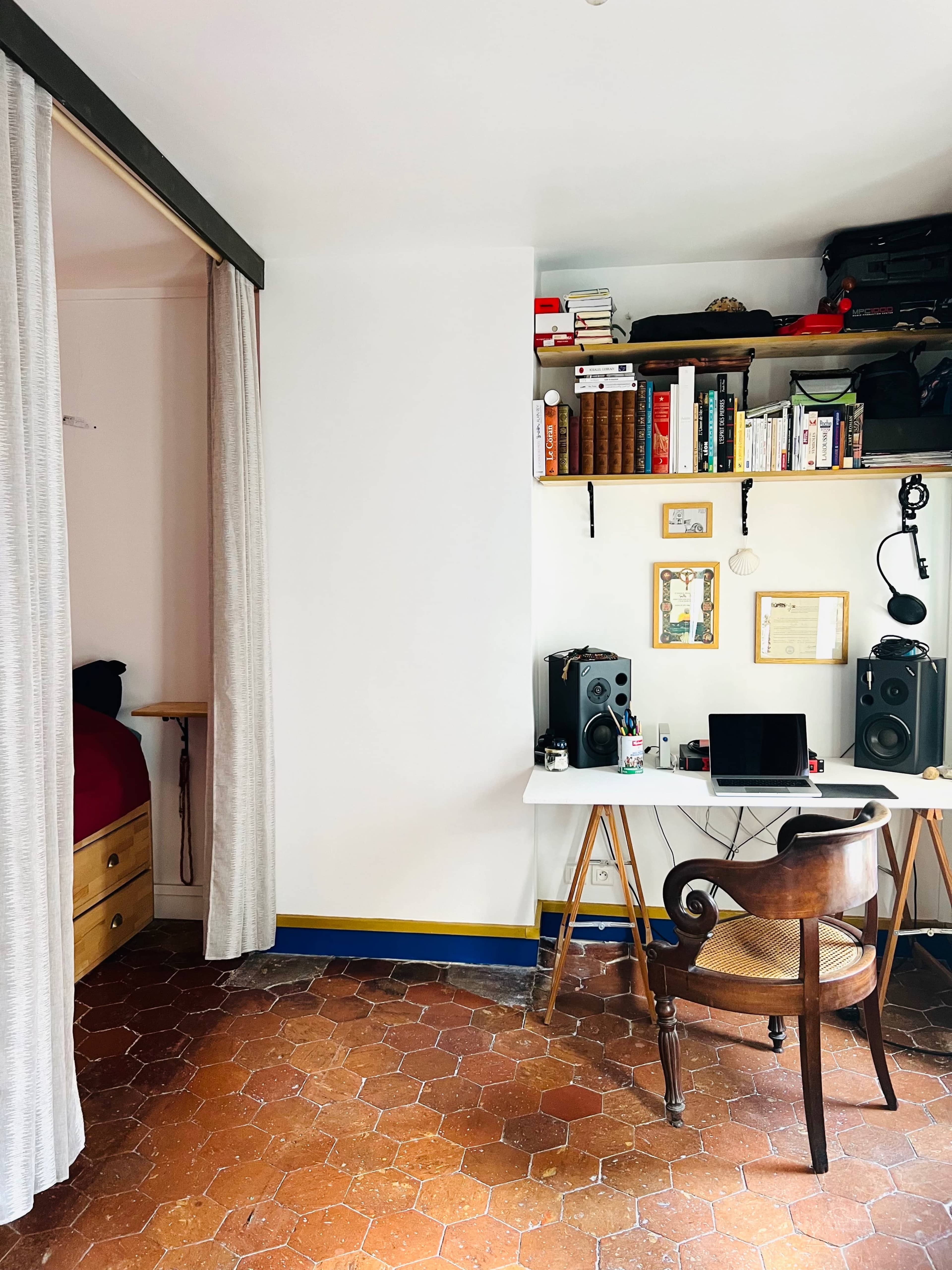 A workspace features a desk with a computer and speakers, surrounded by bookshelves and a curtained area leading to a bed.