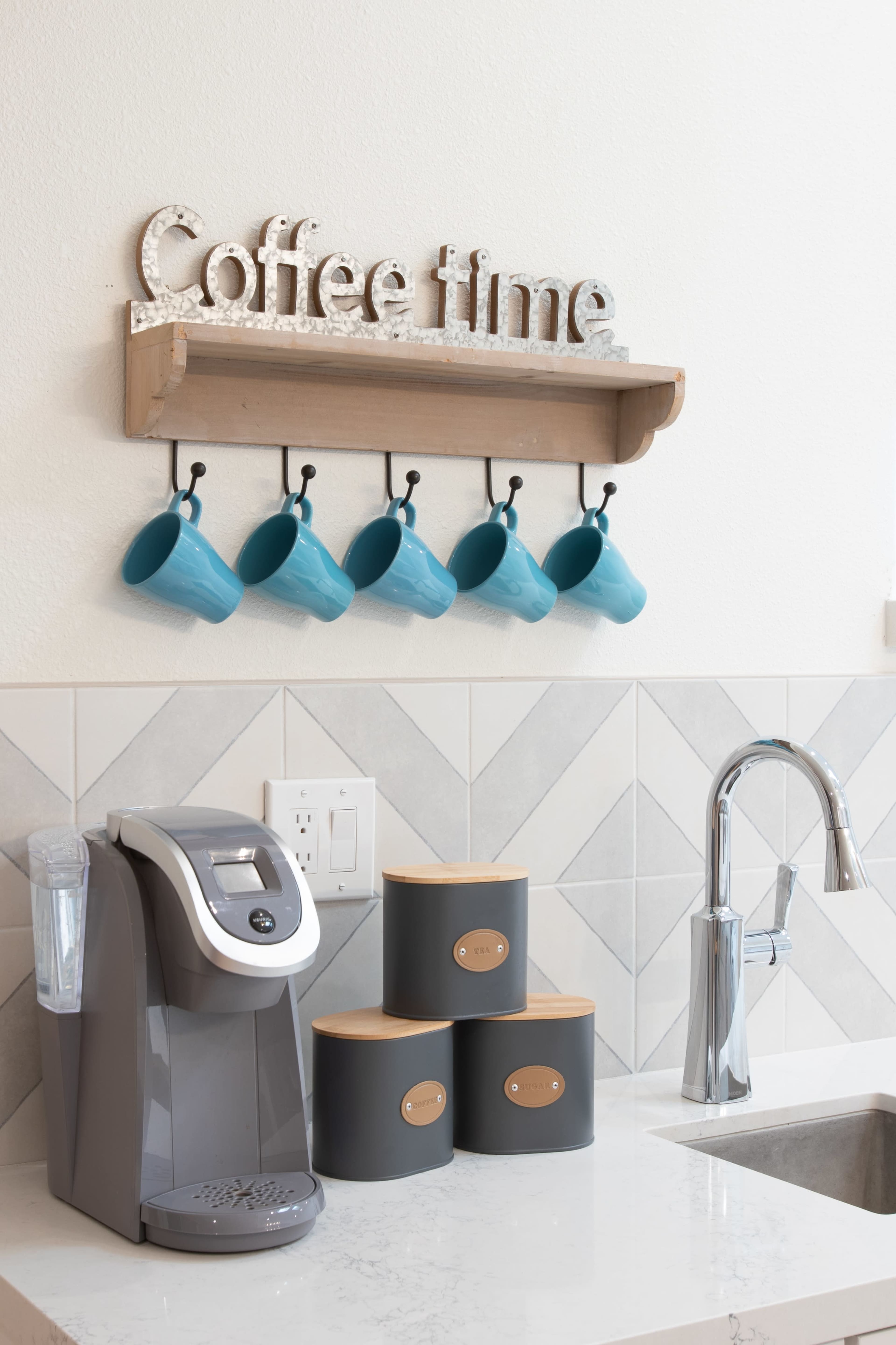 A coffee station features a shelf with the words "Coffee time," five blue mugs hanging below it, a coffee maker, and three canisters on a countertop.
