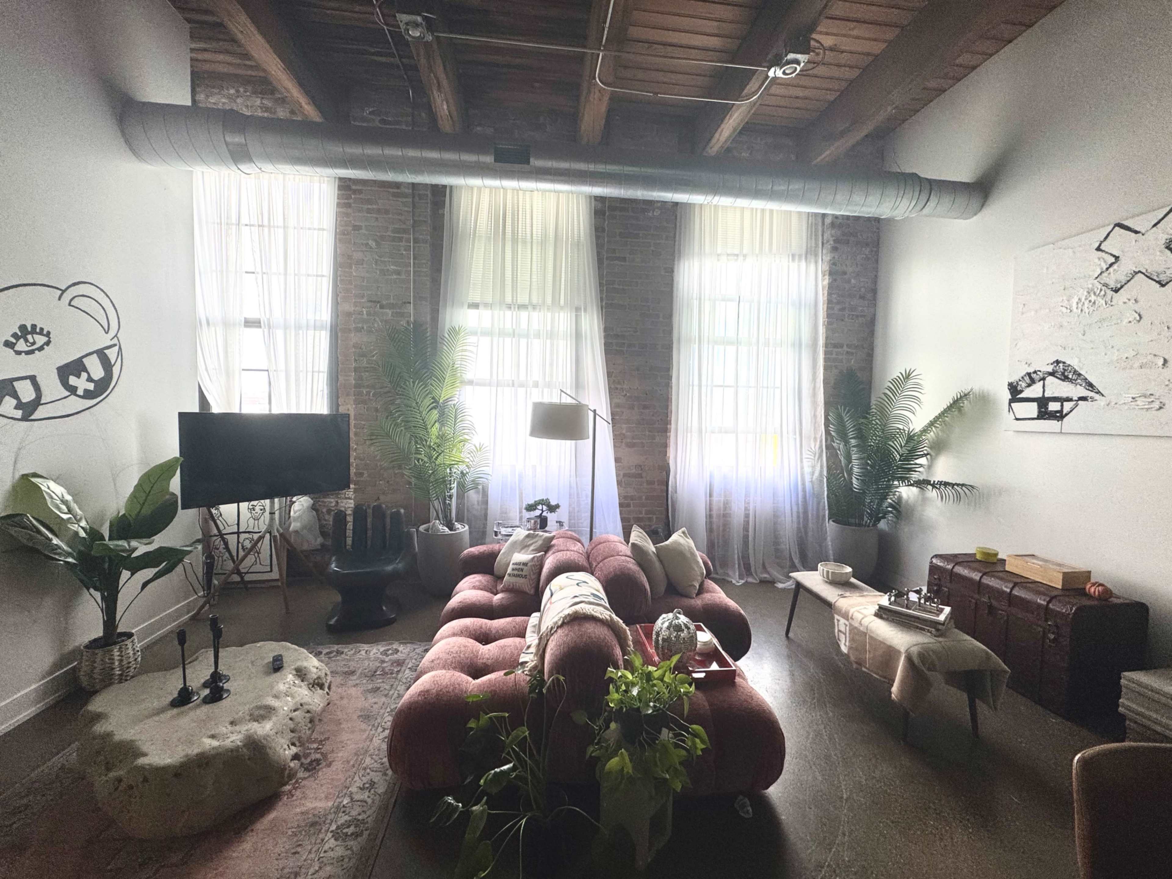 The image shows a spacious living room with large windows, a red tufted sofa, and a mix of plants and decorative elements.