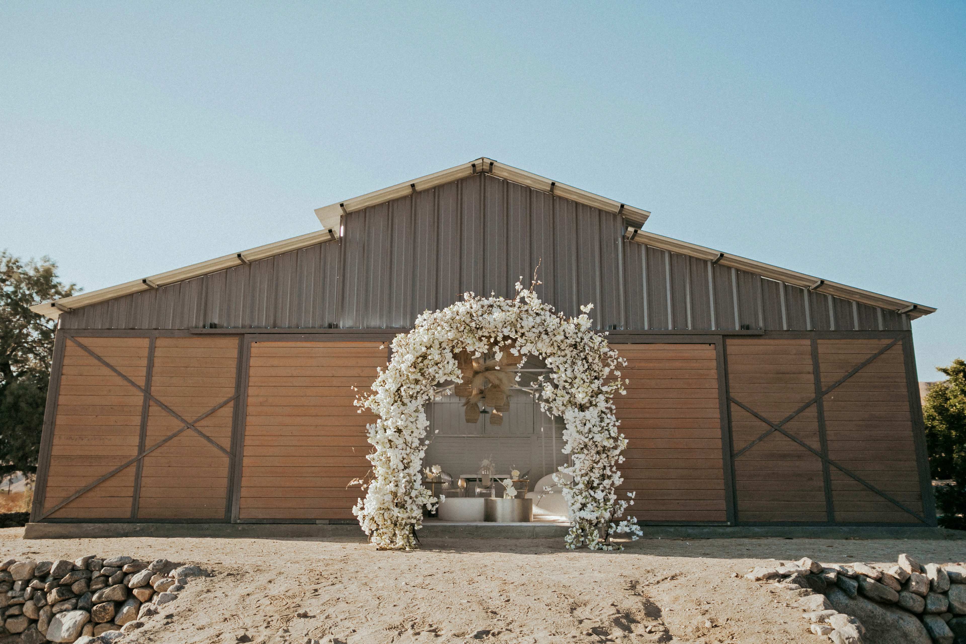 A rustic barn with a floral archway at the entrance is set against a clear blue sky.