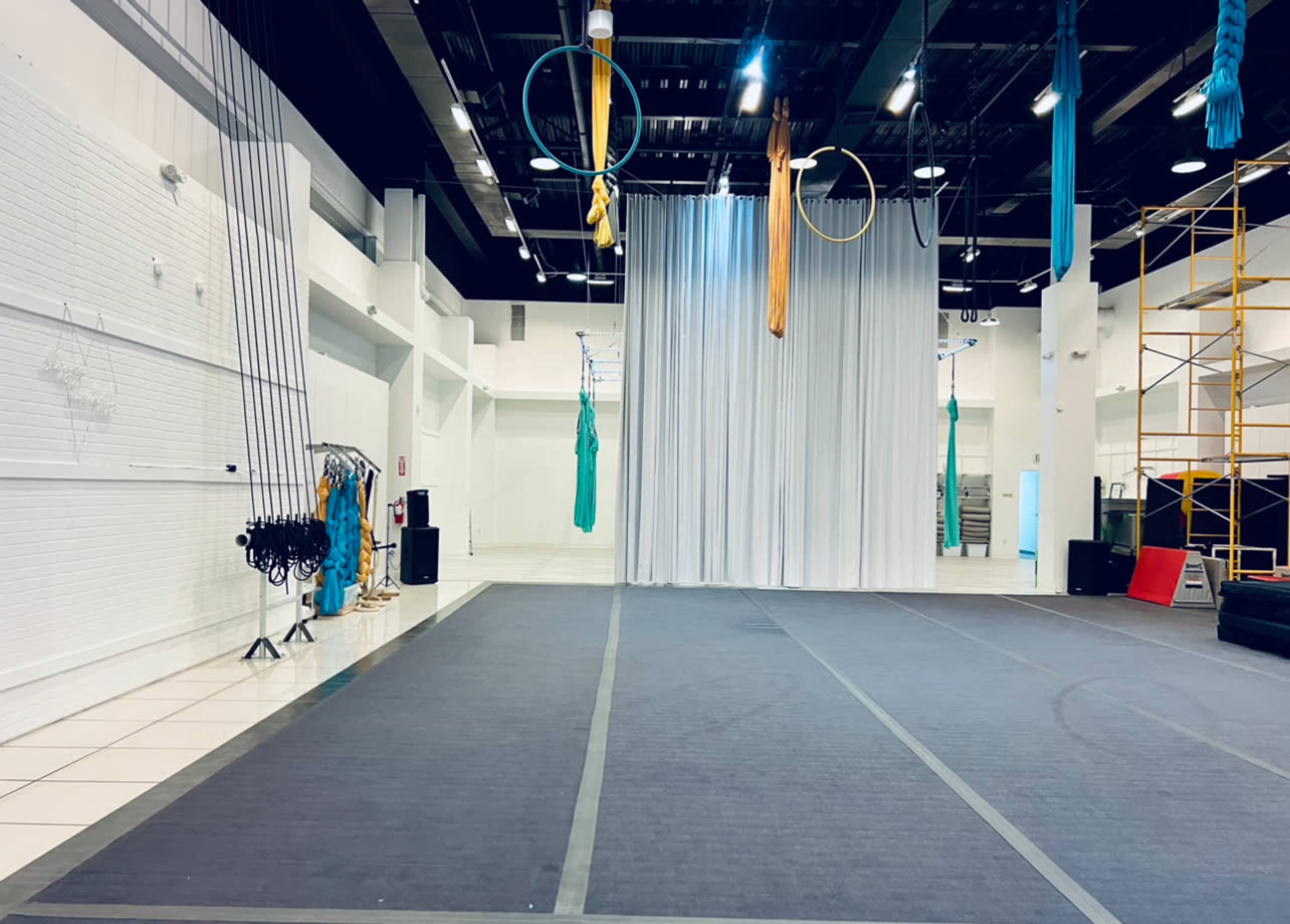 The image shows a spacious indoor area set up for aerial arts, featuring silks, hoops, and a tall fabric curtain, along with equipment like a scaffolding and musical speakers.