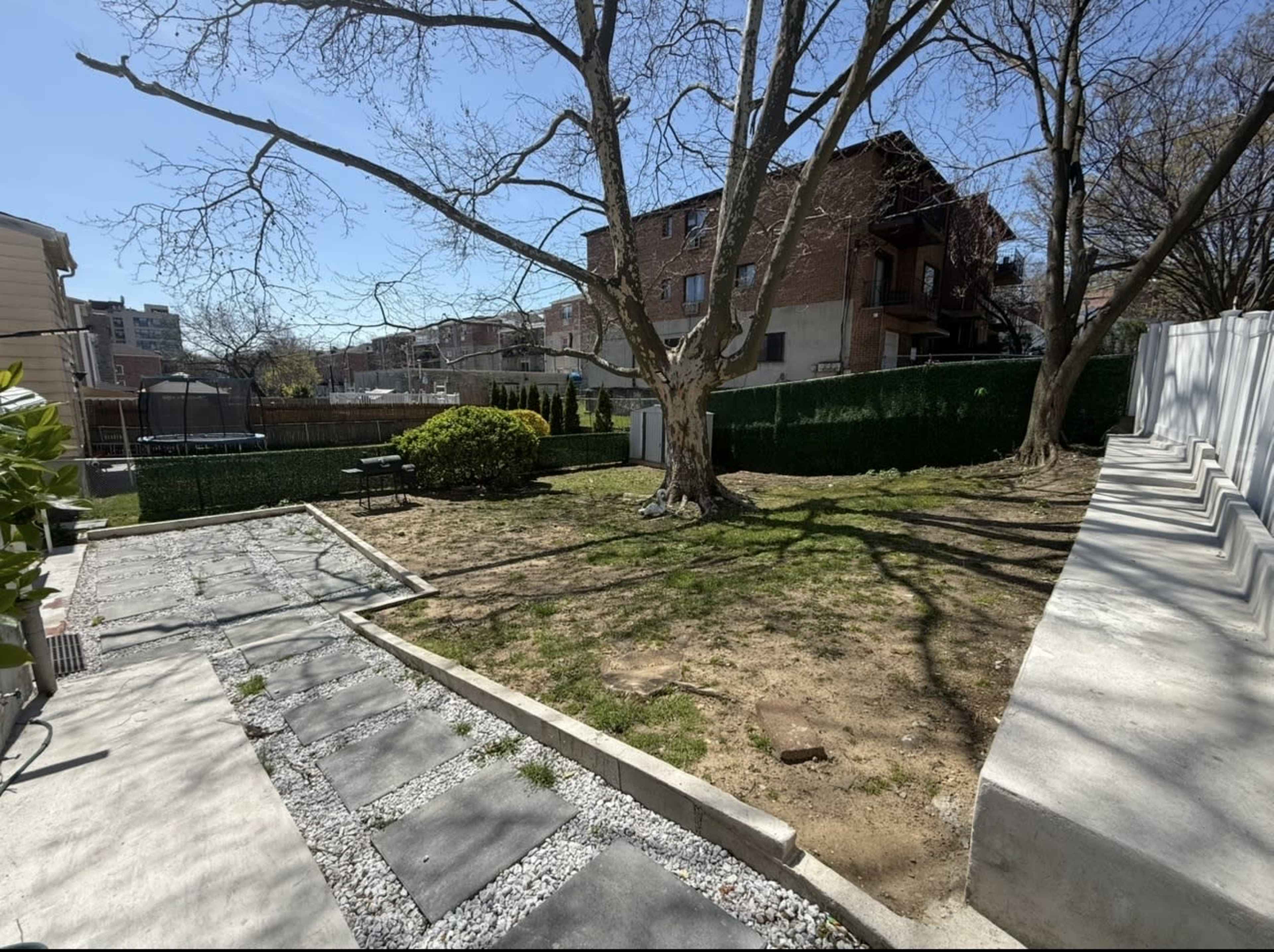 The image shows a sunny backyard with a tree, a patch of grass, a stone walkway, and a fenced area in the background.