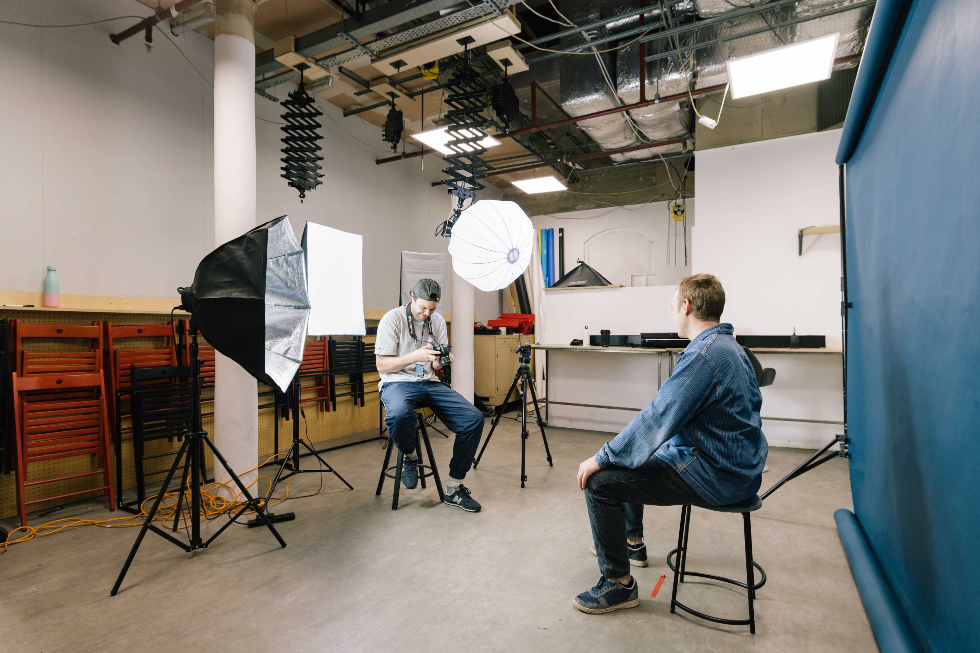 A photographer is setting up equipment in a studio while a subject sits on a stool in front of him.