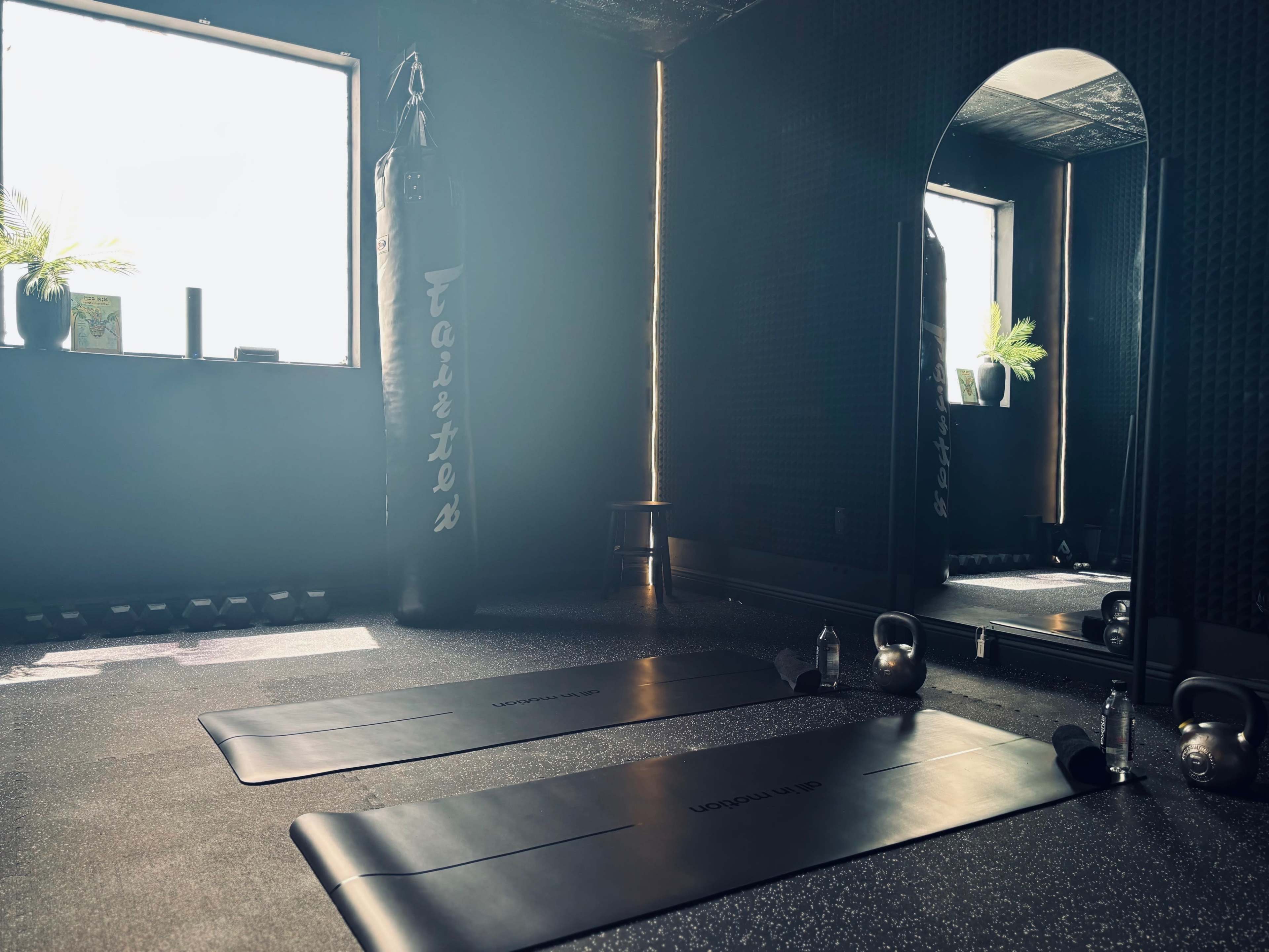 An indoor workout space featuring two exercise mats, a punching bag, a mirror, and kettlebells, with natural light coming through a window.