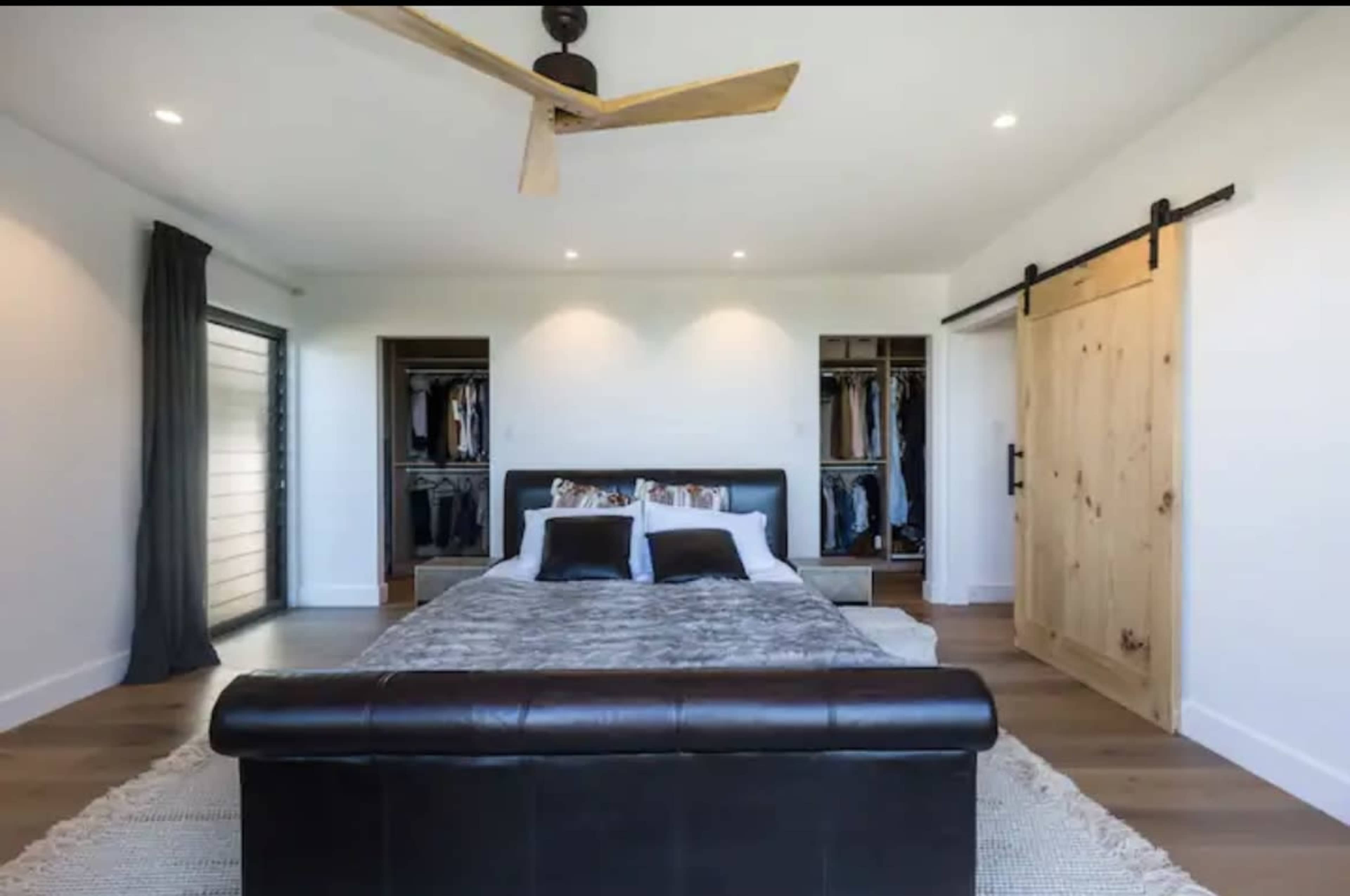 The bedroom features a large bed with a dark leather bench at the foot, a ceiling fan, and a sliding wooden door leading to a closet.