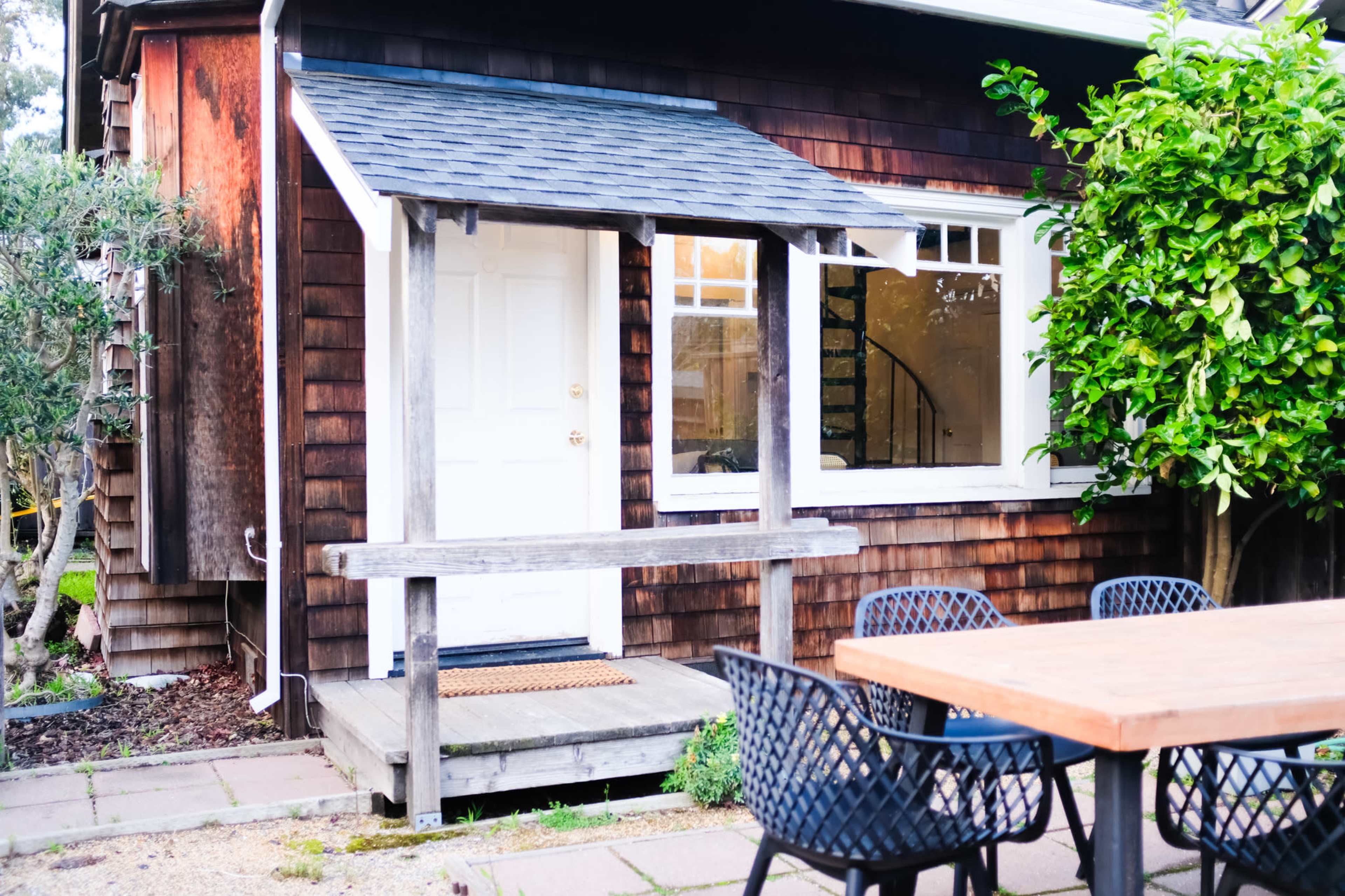 A small wooden porch with a white door, adjacent to a building with dark wooden siding, and a table with black chairs positioned on a patio.
