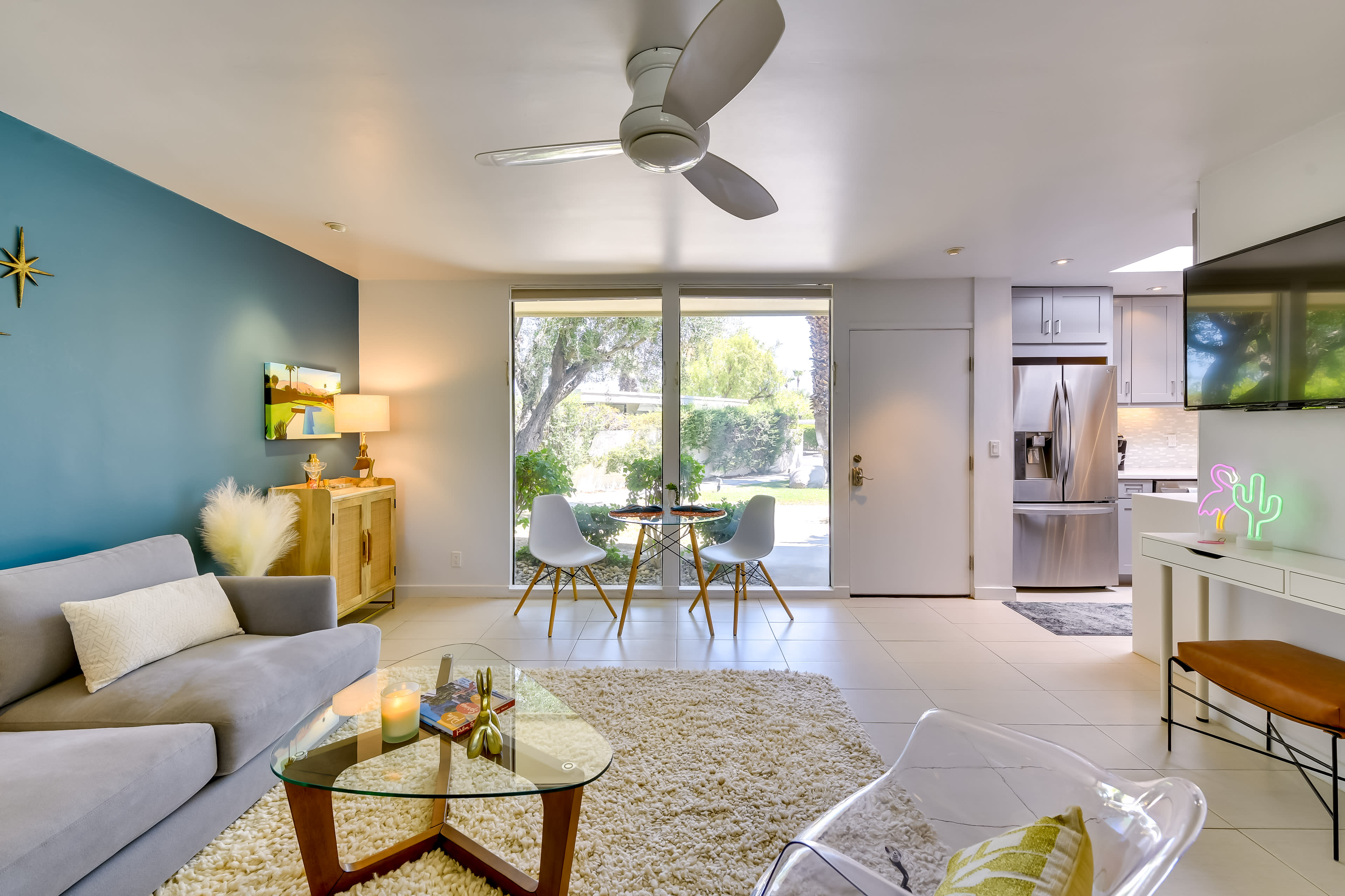 The image shows a modern living room with a gray sofa, a glass coffee table, and a dining area by a window, leading to an outdoor view.
