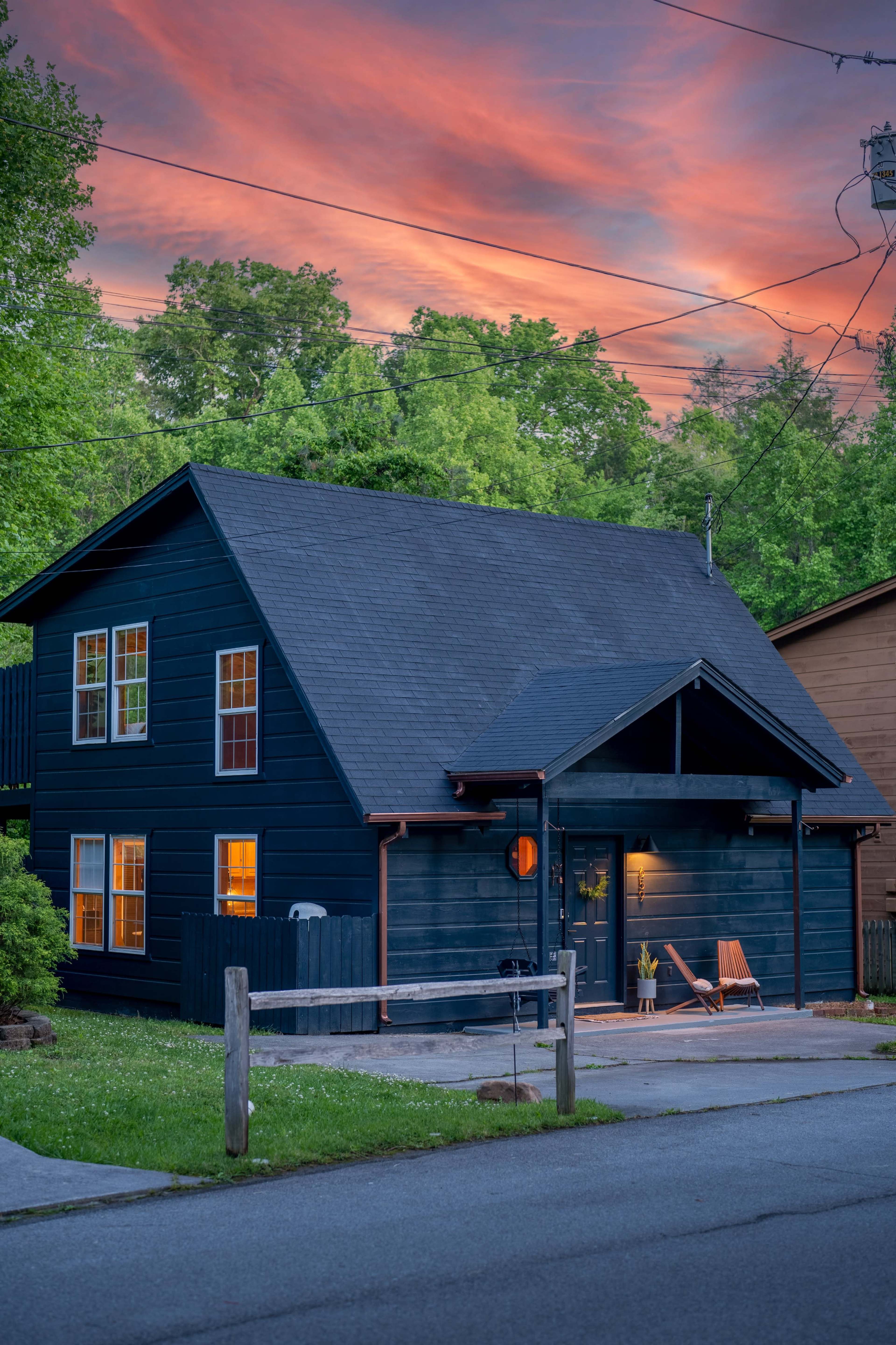 A dark-painted house with a sloped roof is situated on a serene street, illuminated by the warm glow of sunset.