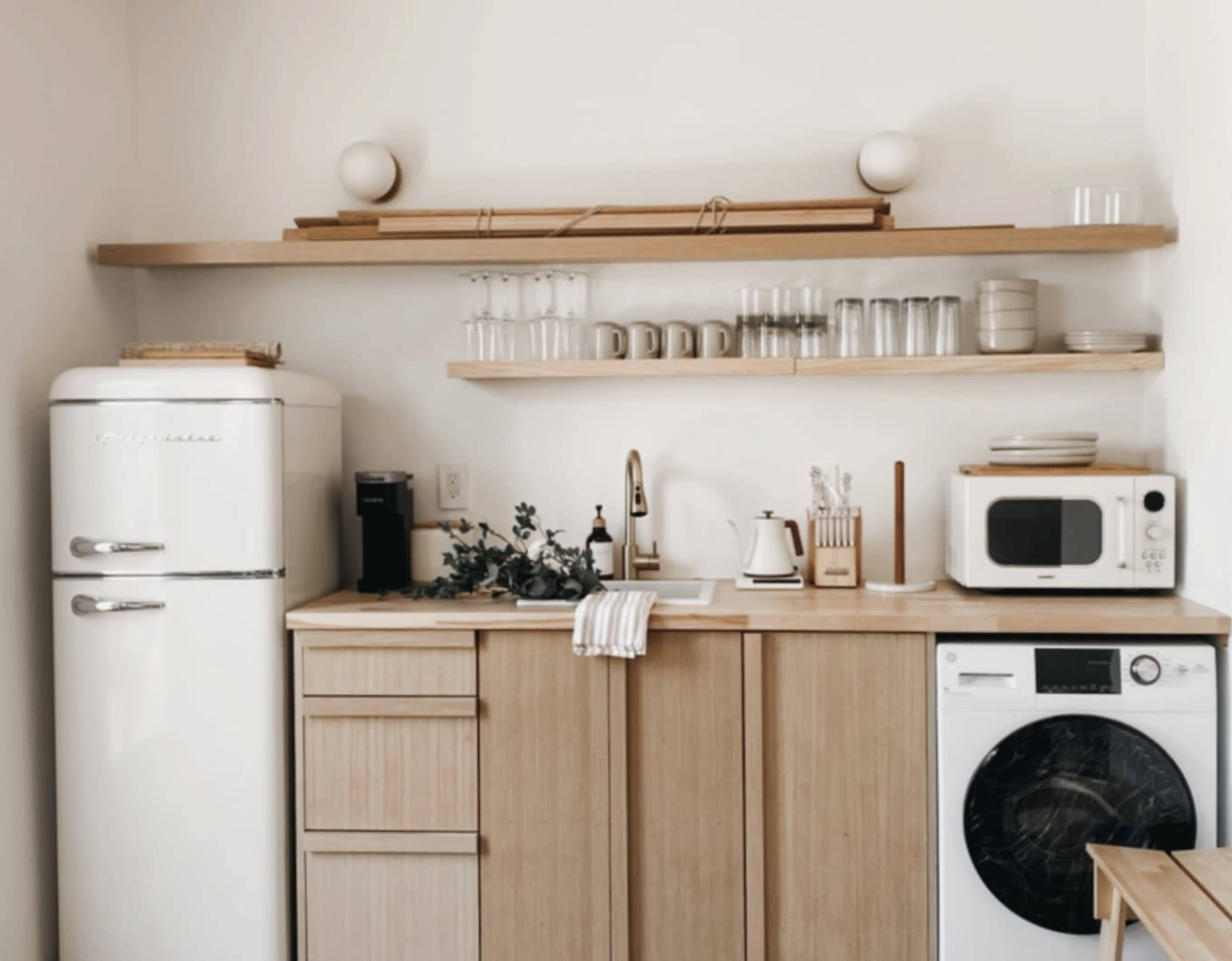 The image shows a modern kitchen with a white retro refrigerator, wooden cabinetry, open shelves displaying glassware, and a washing machine.