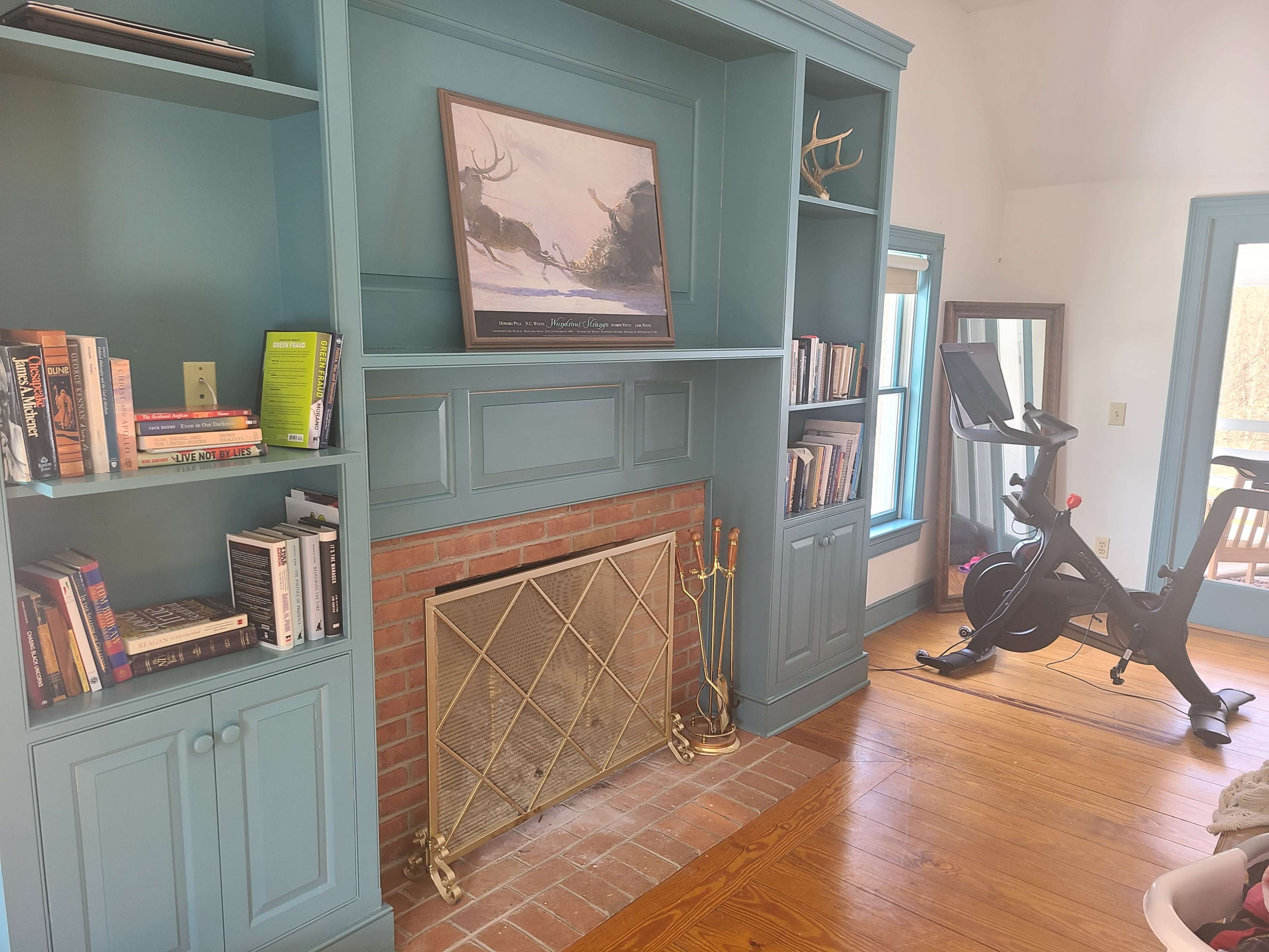 The image shows a room with a built-in bookshelf, a brick fireplace, a mounted painting, and an exercise bike.