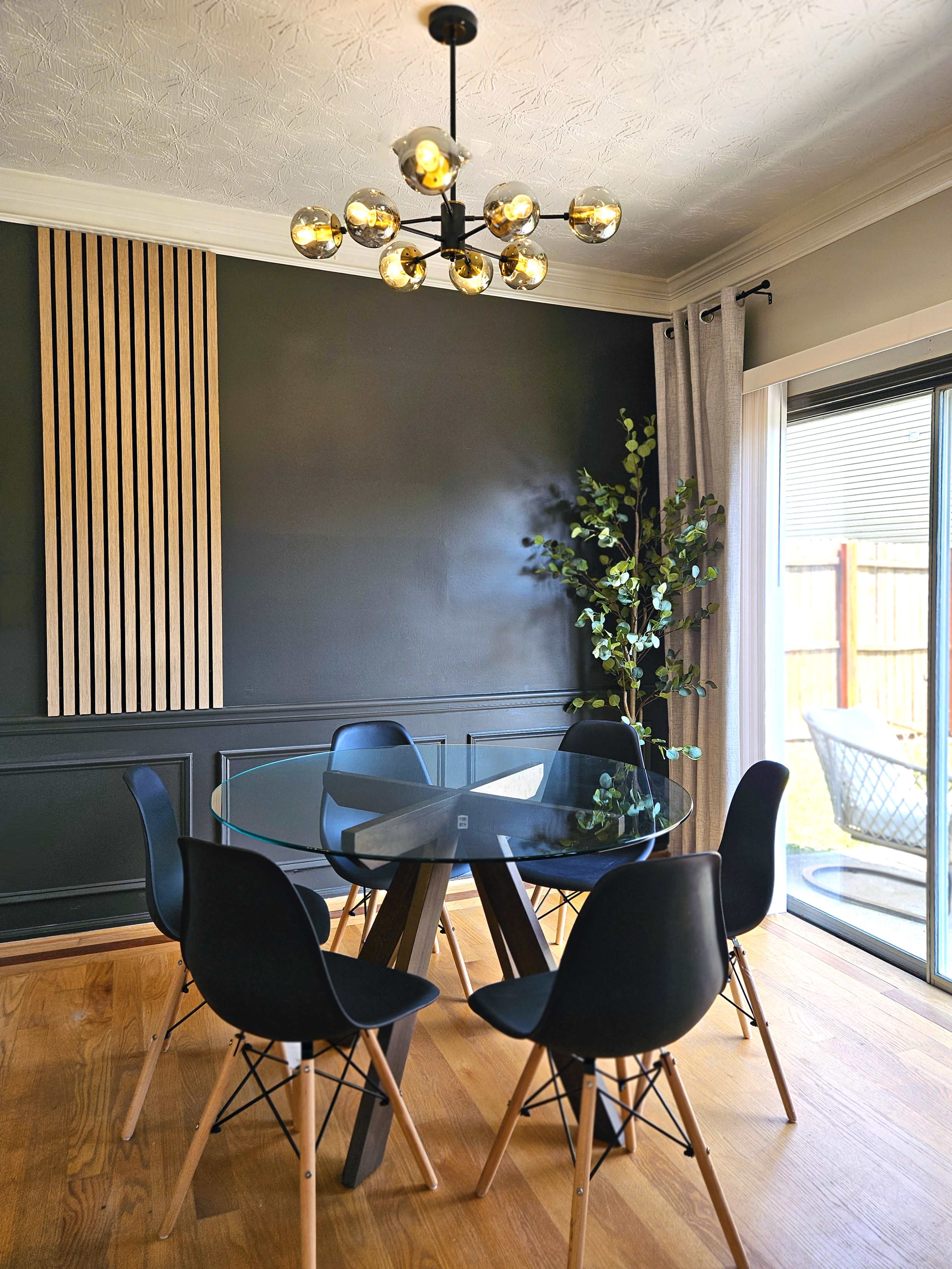 The image shows a modern dining area with a glass table surrounded by black chairs and a decorative light fixture above, complemented by indoor plants and wooden accents on the wall.