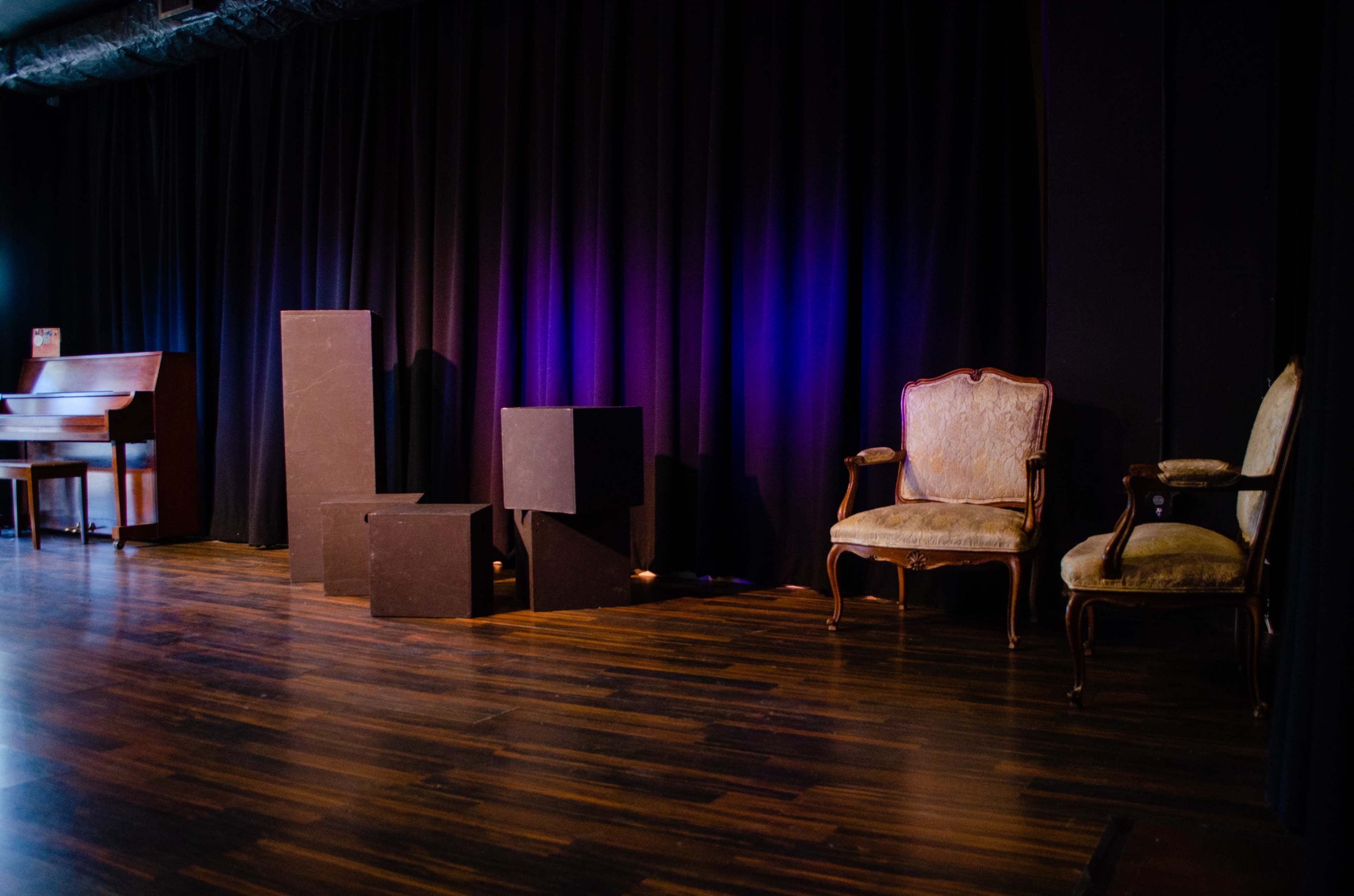A stage is set with a black curtain in the background, a vintage chair on one side, and various black geometric shapes scattered across the wooden floor.