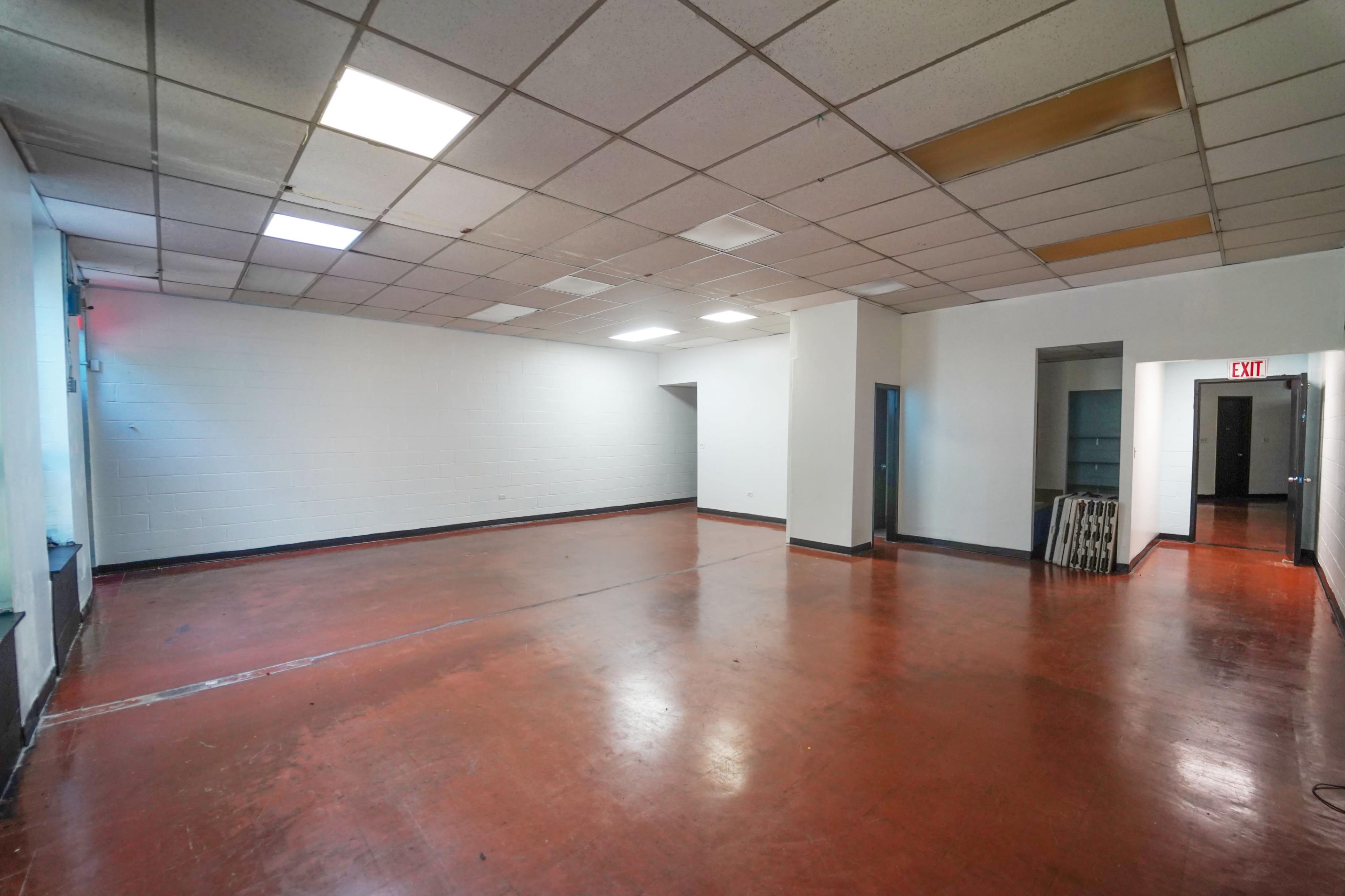 The image shows a large, empty room with a red concrete floor, white walls, and fluorescent ceiling lights.