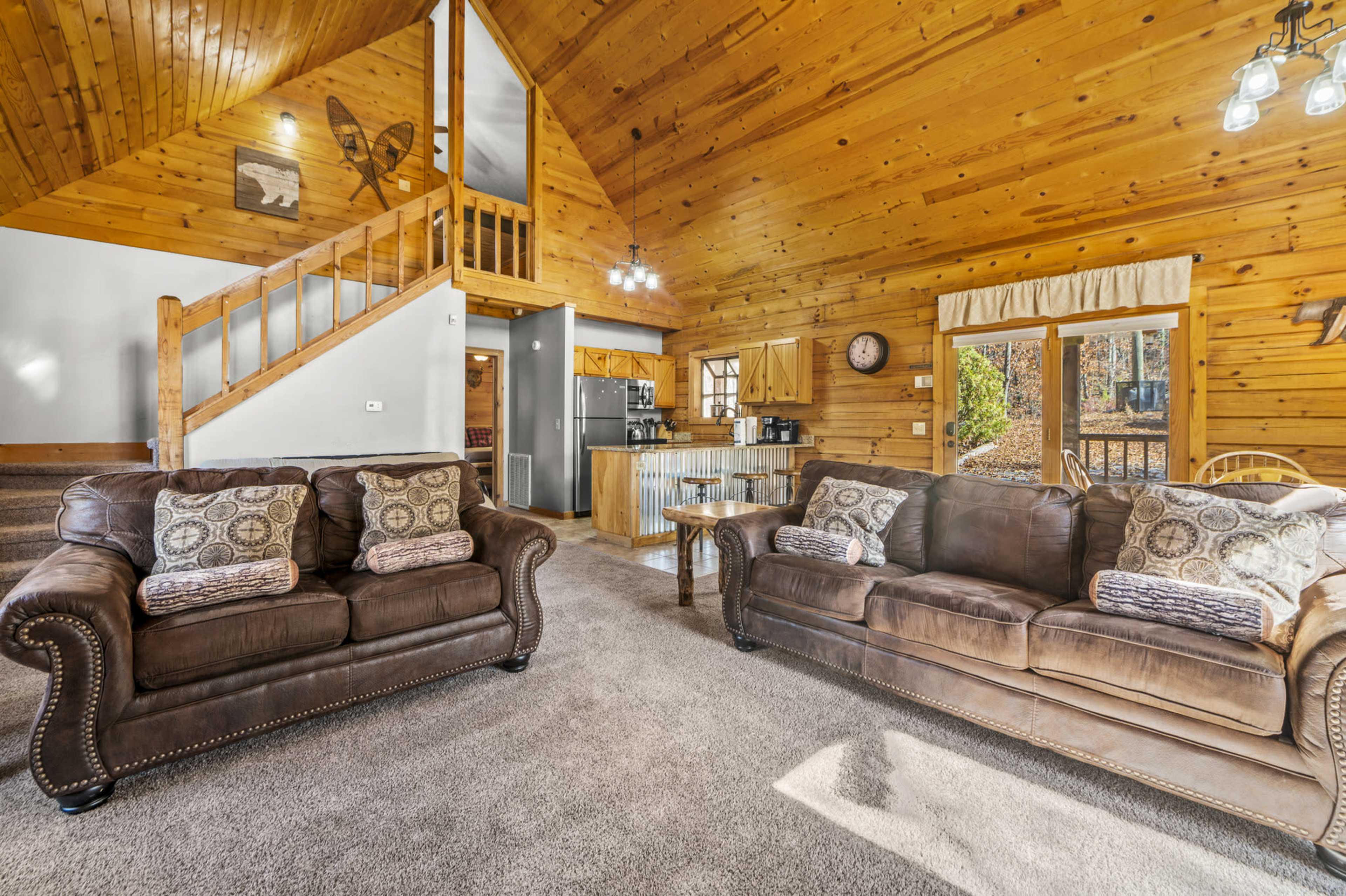 The interior of a wooden cabin features two brown sofas in a cozy living area with a staircase leading to an upper floor and a kitchen visible in the background.