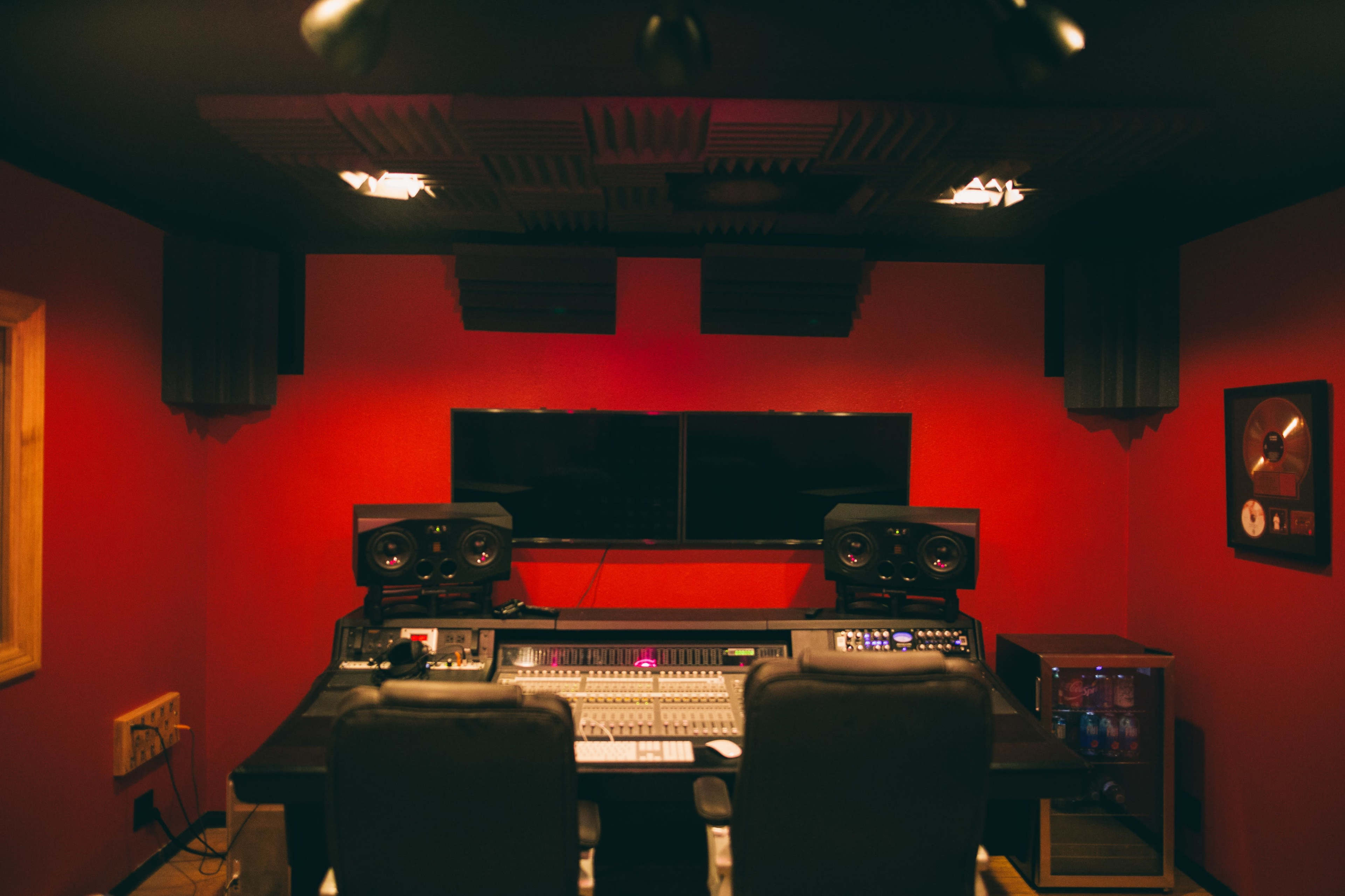The image shows a recording studio with a black mixing console, two large speakers, and sound-absorbing panels on red walls.
