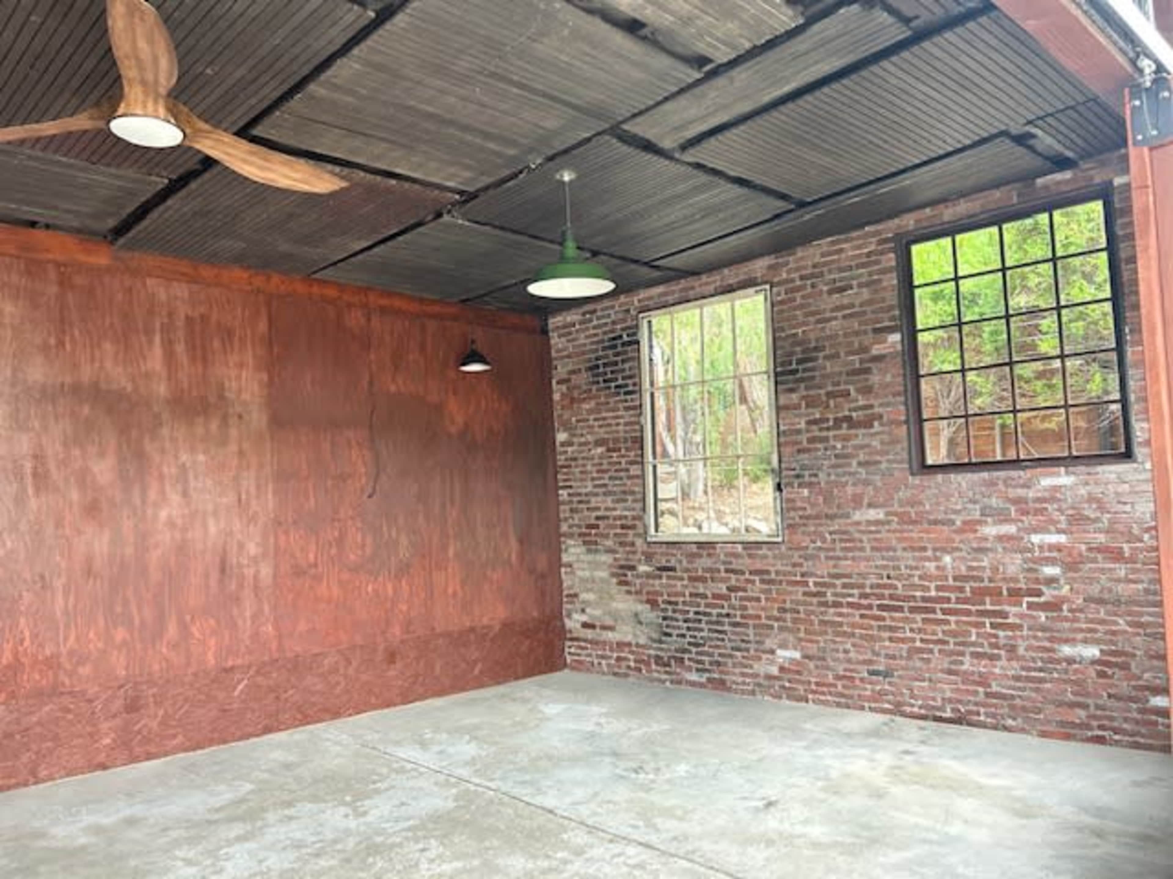 An unfinished interior space with exposed brick walls, a wooden accent wall, a ceiling fan, and two windows.